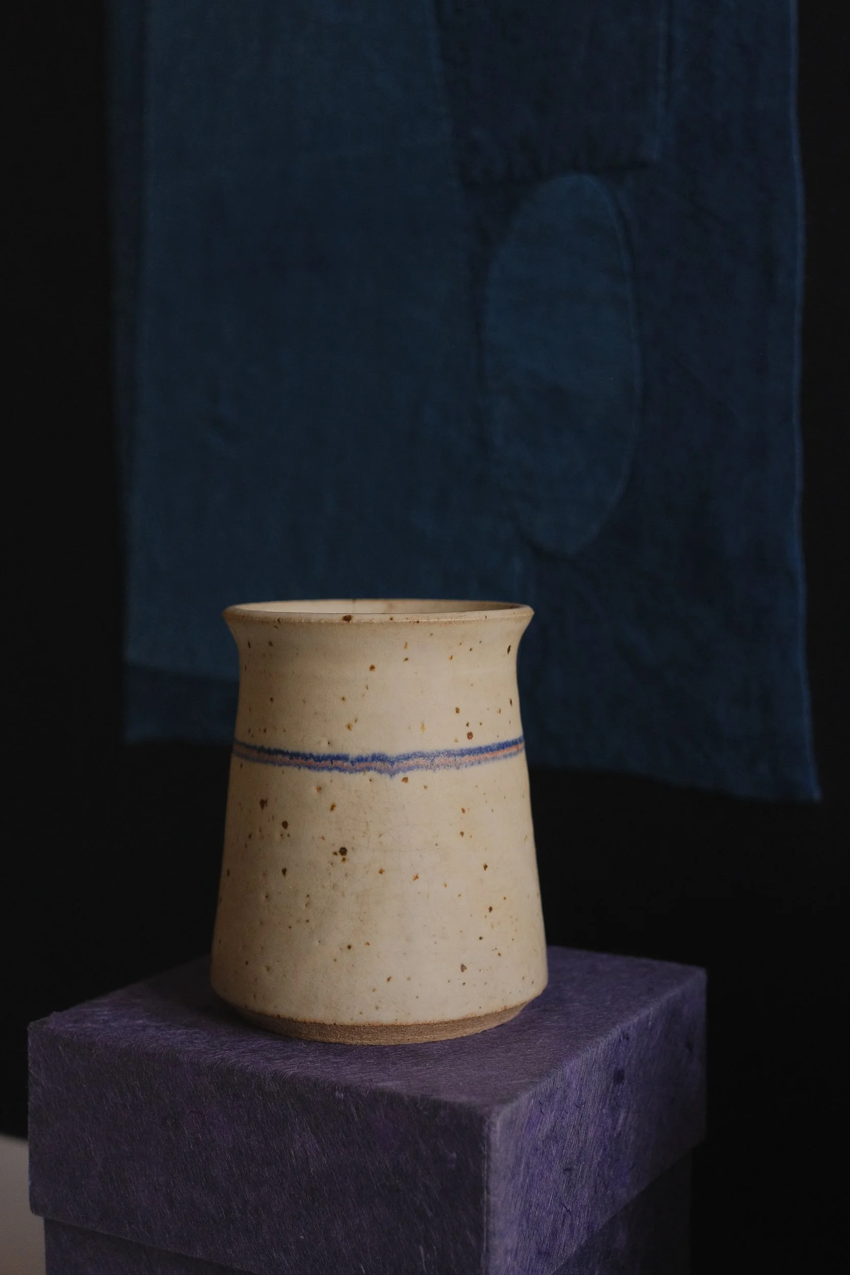 A handmade ceramic cup with a blue line on a purple block, against a dark background with a textured blue cloth hanging behind.