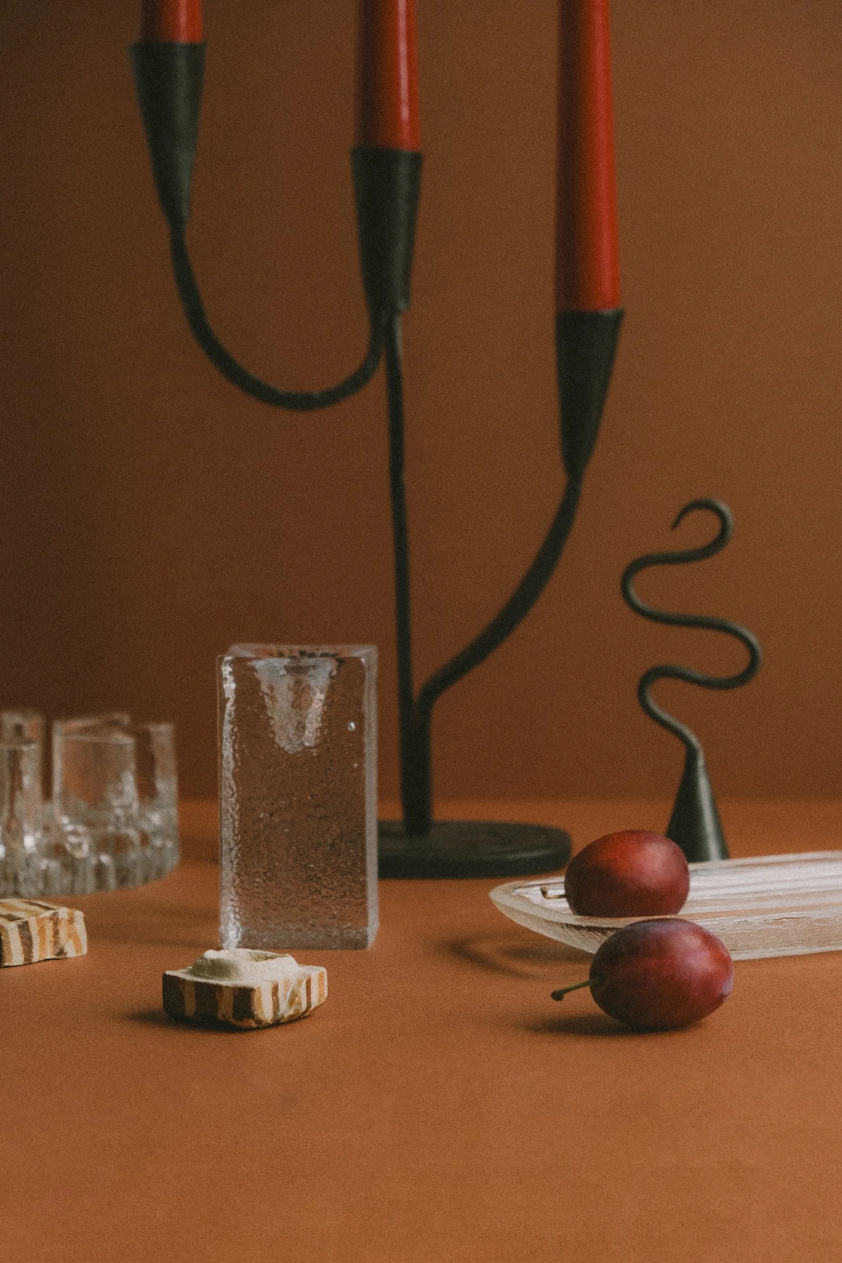 Decorative still life with a black and red candelabrum, a textured glass block, two red plums on a white dish, and small glass containers on a wooden surface against a brown background.