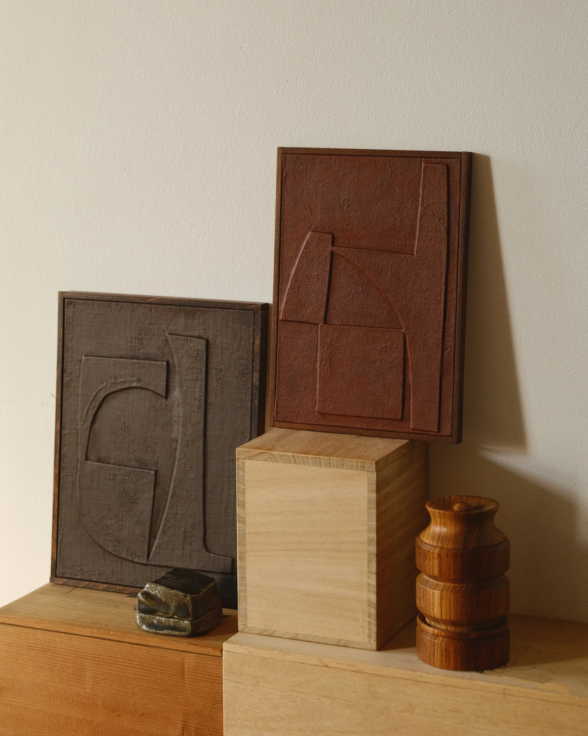 Display of two carved wooden blocks with geometric shapes, one dark gray and one red, placed on wooden blocks with small wooden and stone objects nearby.