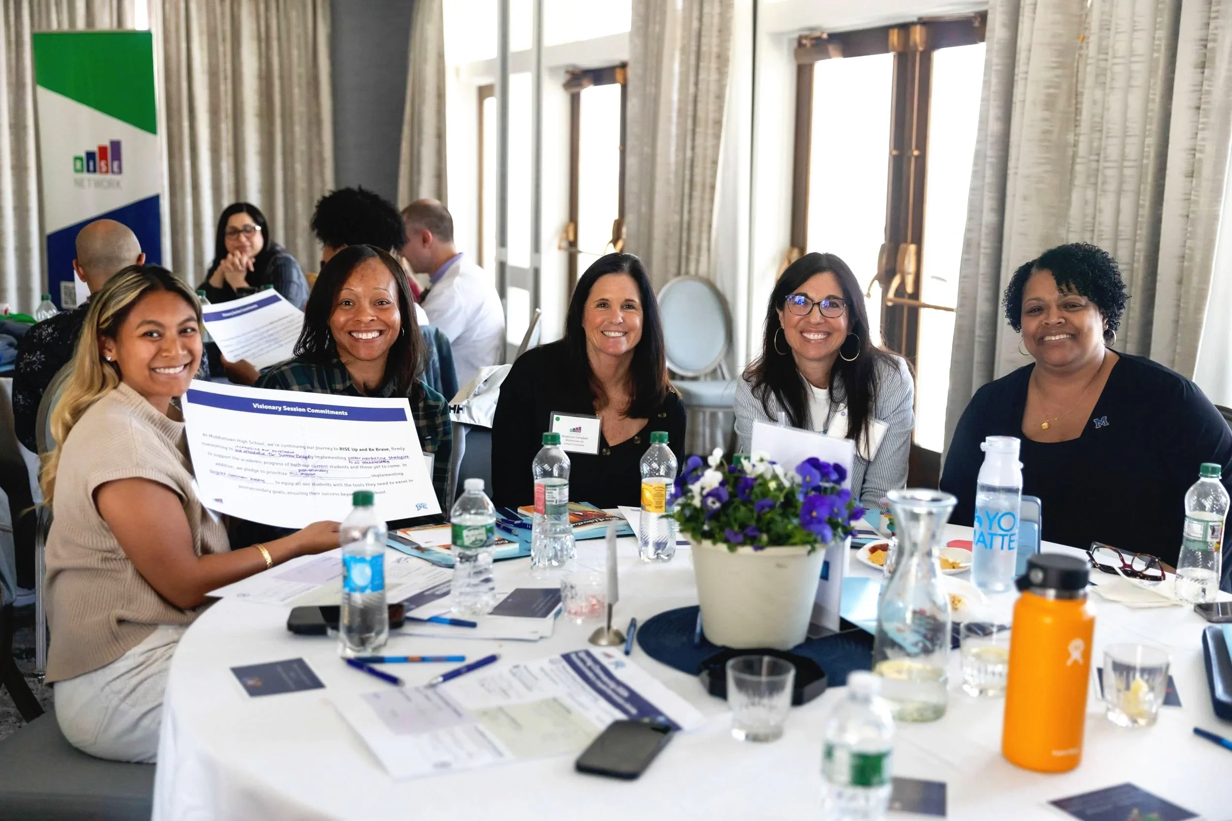 Participants at a nonprofit workshop smiling and holding documents while seated around a round table during a collaborative session.