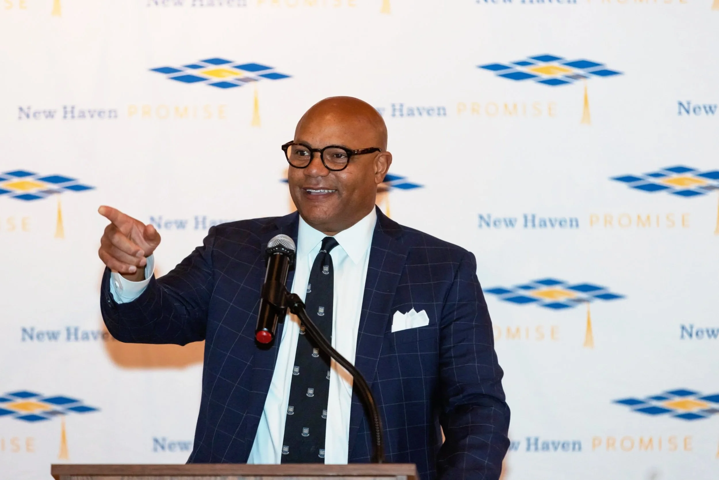 Nonprofit executive speaking at a podium during a New Haven community event with organizational branding behind him.