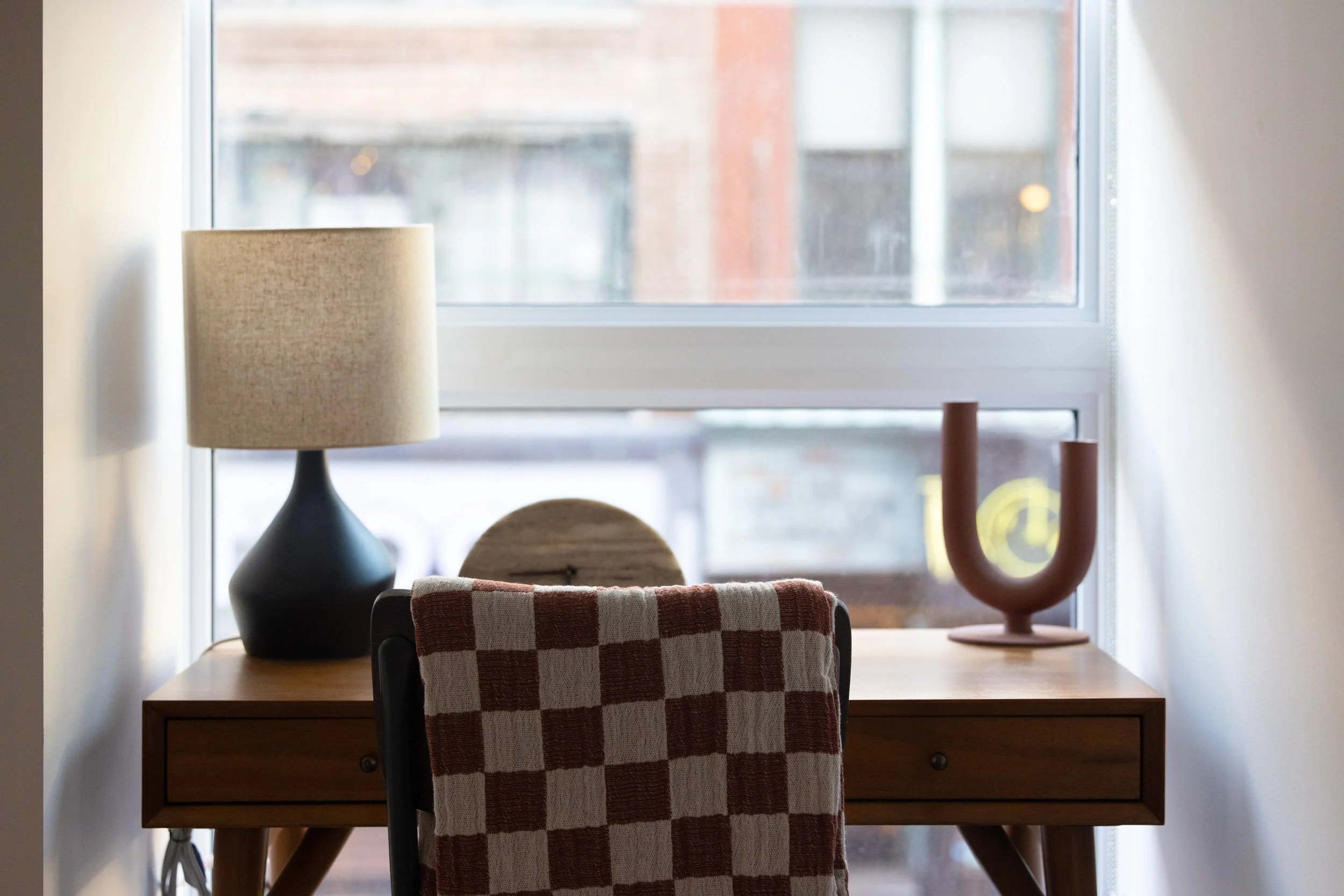 Styled residential workspace with wooden desk, table lamp, and natural light from large window in Connecticut apartment