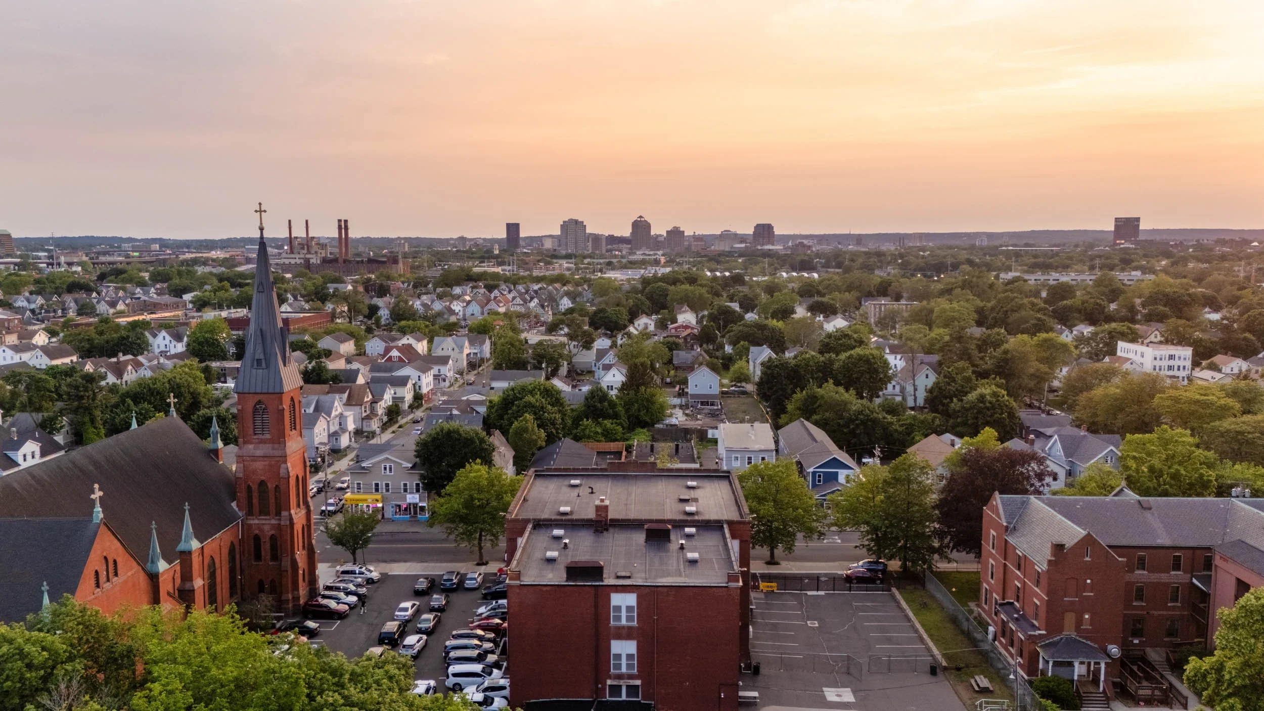 Aerial drone view of a residential neighborhood in New Haven, Connecticut, with a church and city skyline visible at sunset.