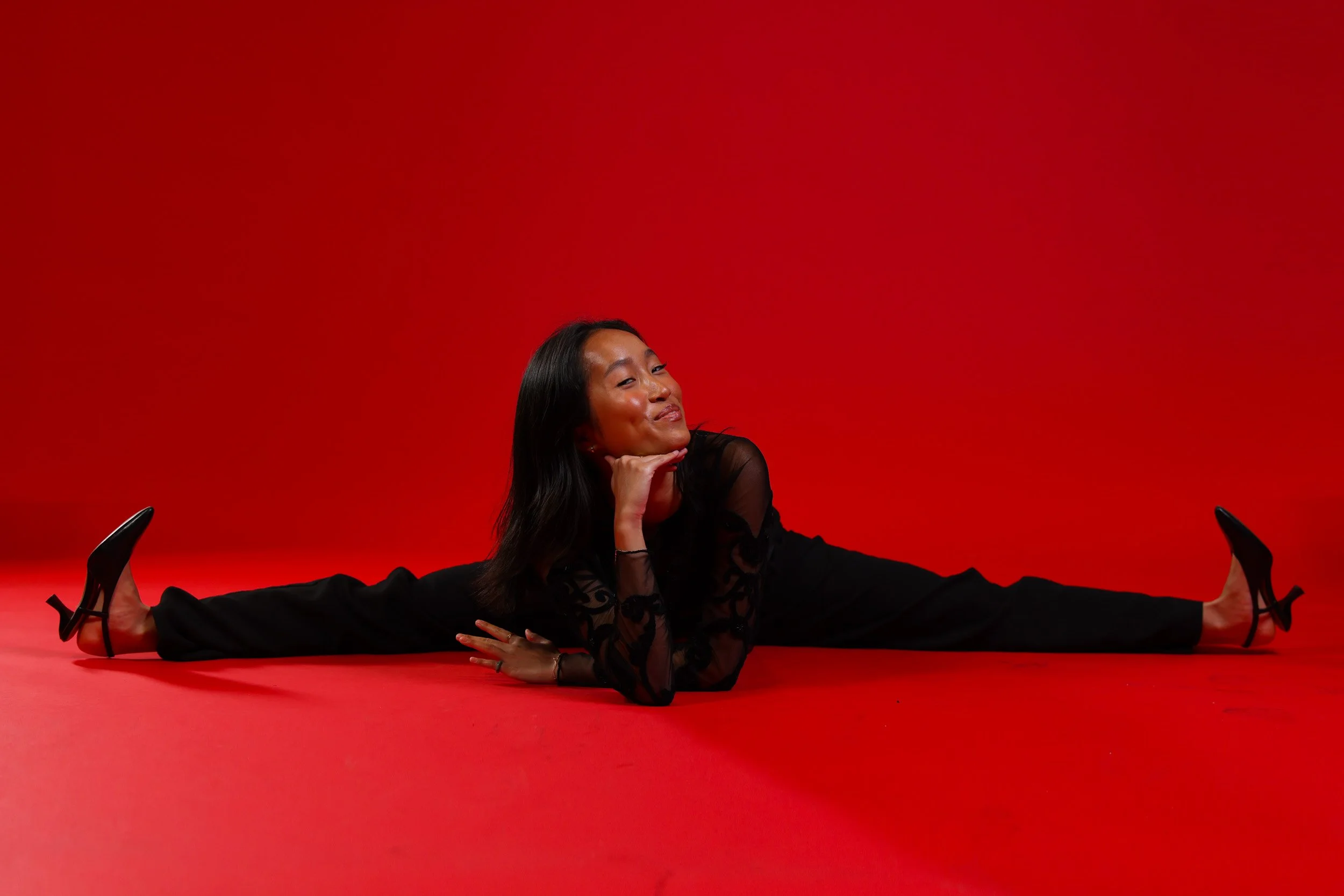 A studio portrait set against a bold red backdrop, emphasizing strength, flexibility, and personality. The dancer’s pose is playful yet controlled, highlighting confidence and range.