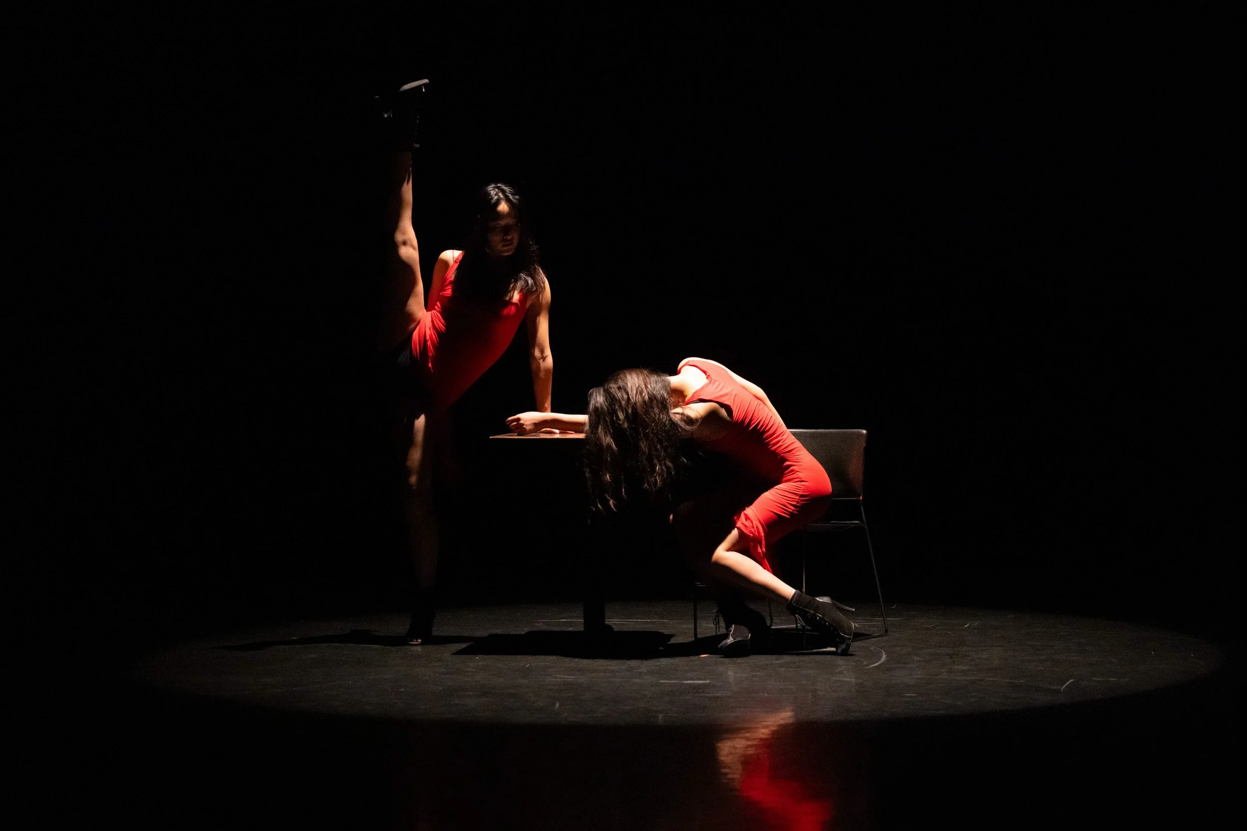 Two dancers in red costumes explore contrast and connection, using chairs as anchors within the choreography. The scene feels intimate and dramatic, shaped by light, posture, and pause.