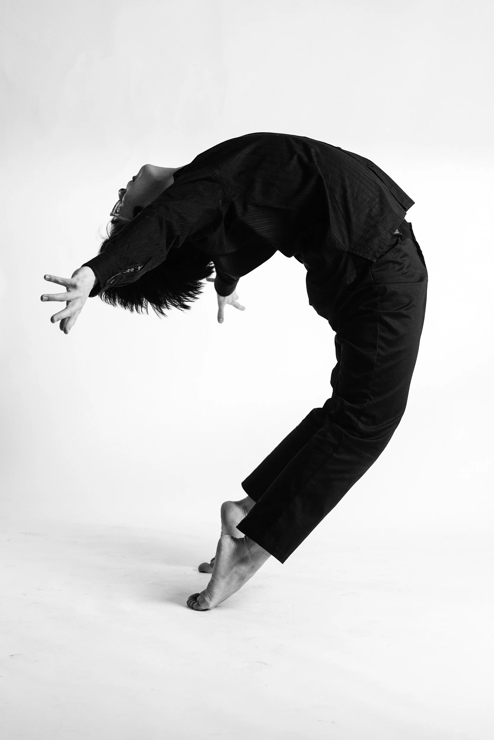 A dramatic backbend unfolds against a white background, emphasizing curve, balance, and breath. The image distills dance to its essential form and physical expression.