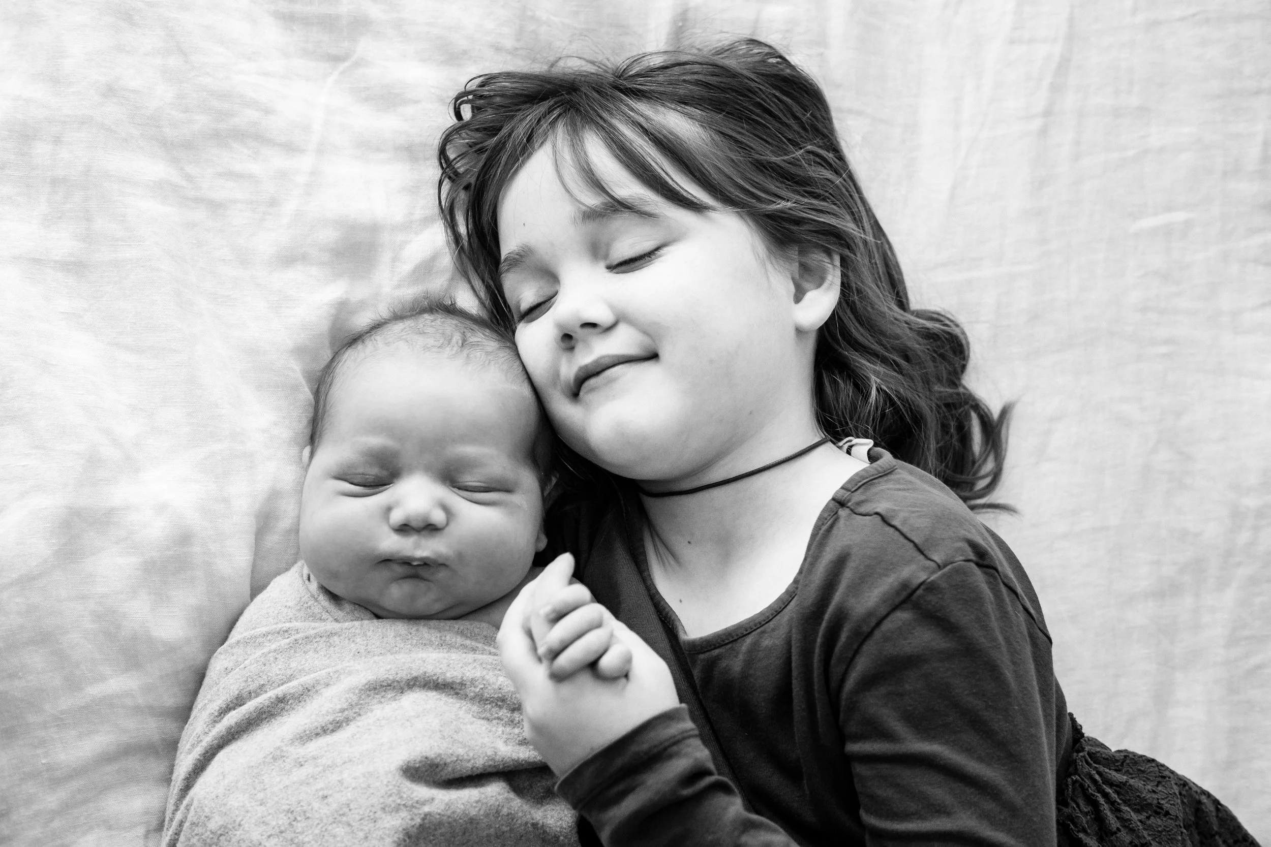Black and white photo of a young girl with closed eyes smiling, cuddling with a baby who also has closed eyes, both resting on a soft surface.