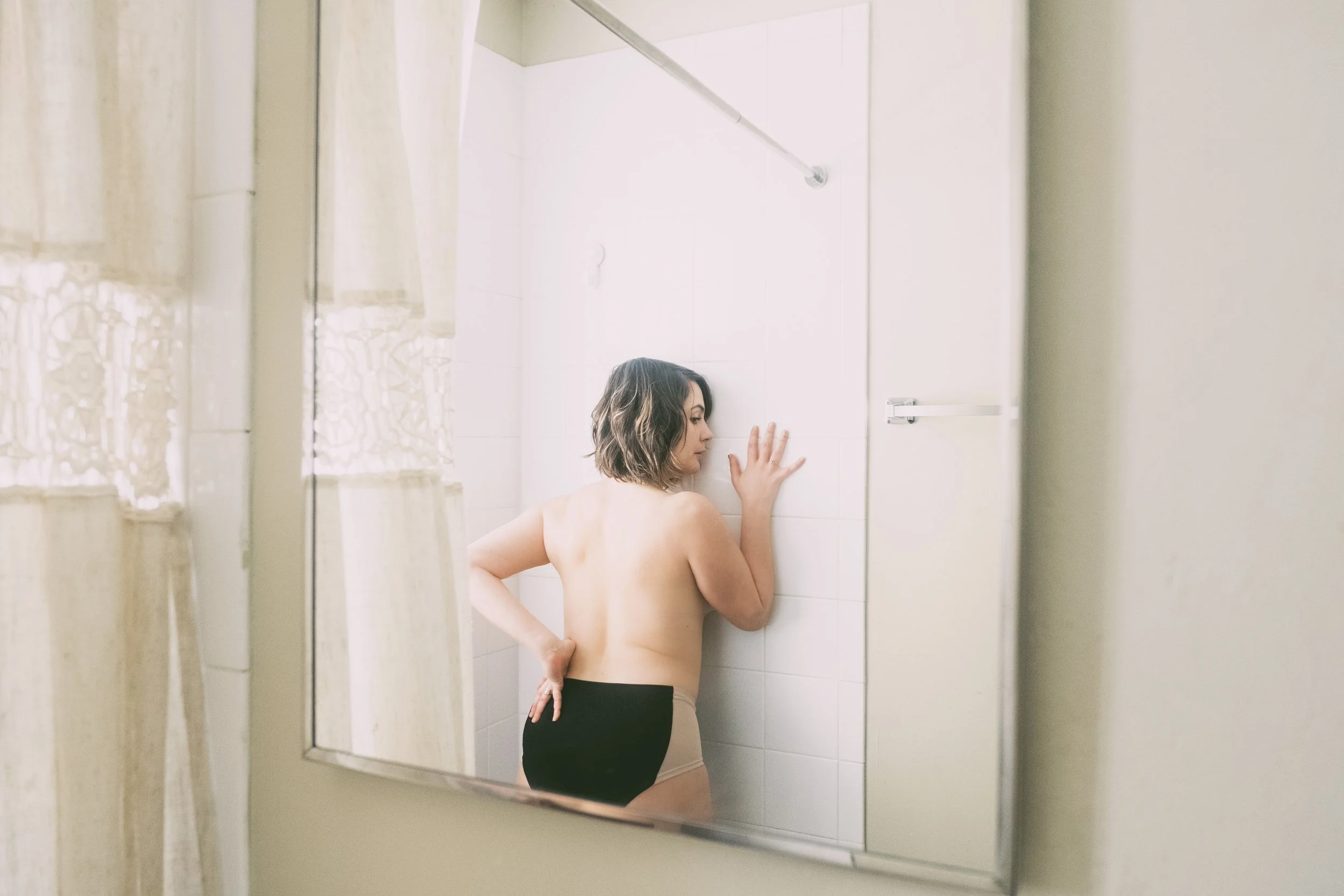 A woman standing in a shower, touching the wall, seen through a bathroom mirror from outside the shower.