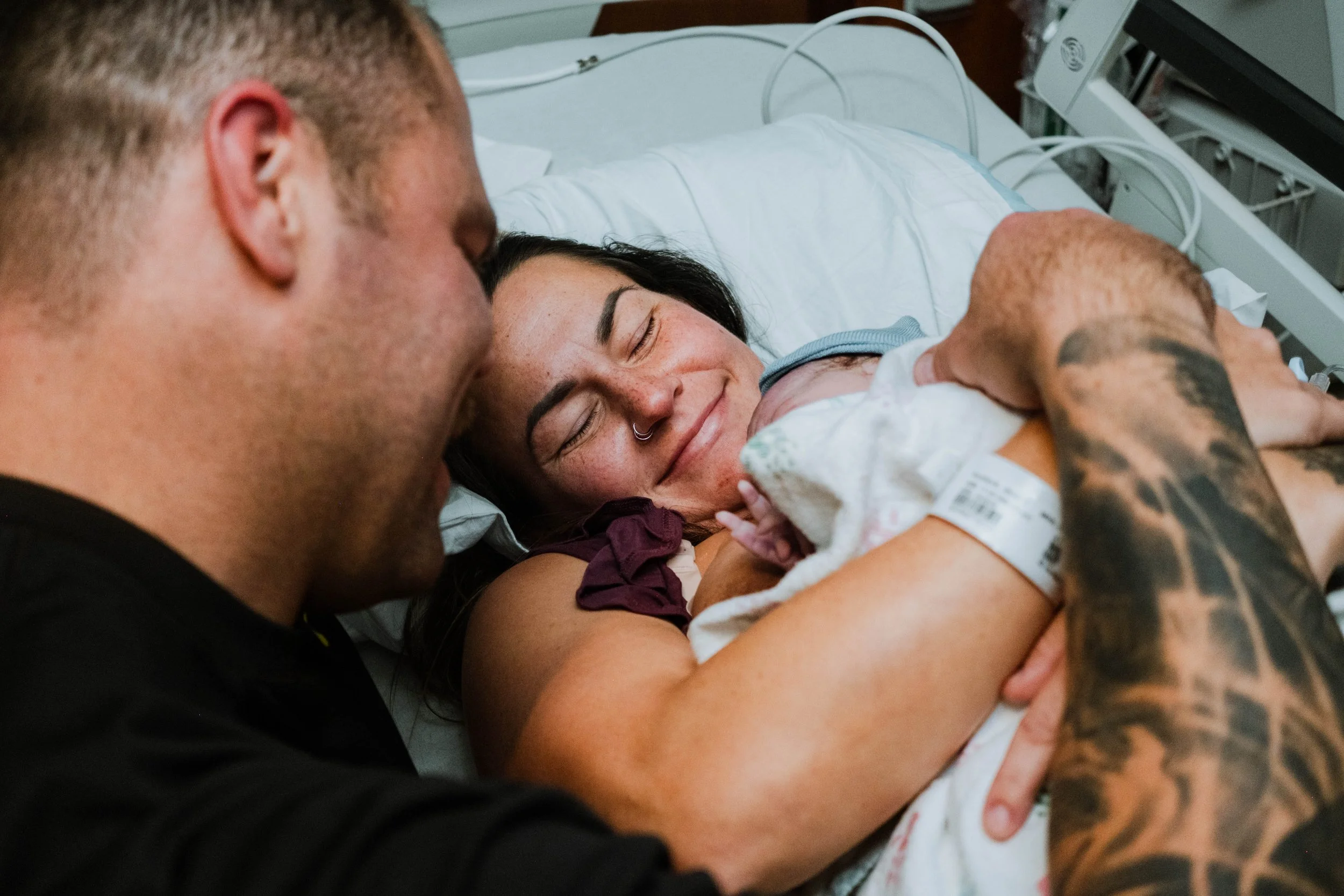 A woman lying in a hospital bed holding a newborn baby, with a man leaning over her, both smiling and looking at the baby.