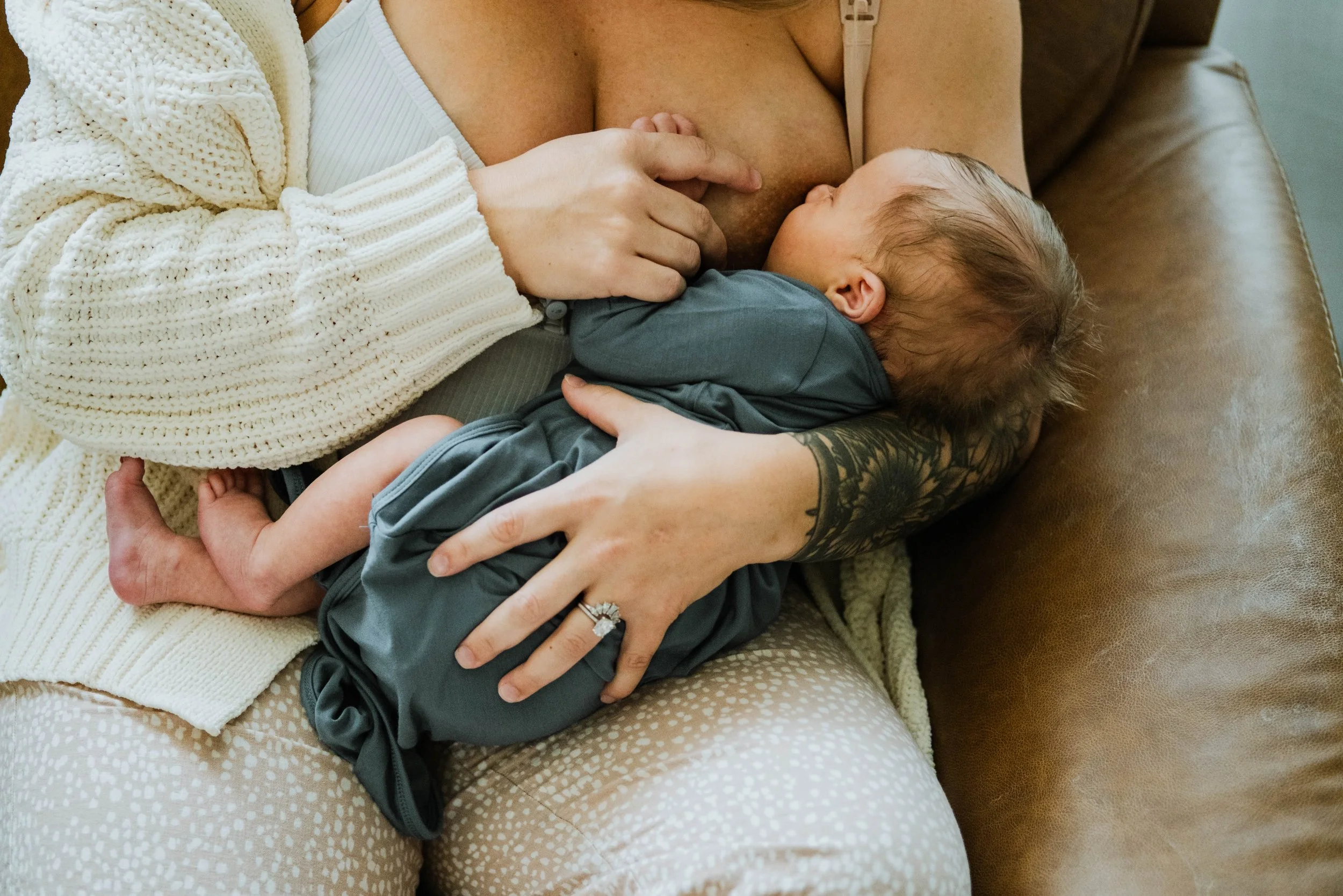 A woman breastfeeding a baby while sitting on a couch, with the woman wearing a creamy knit cardigan and the baby in gray clothing.