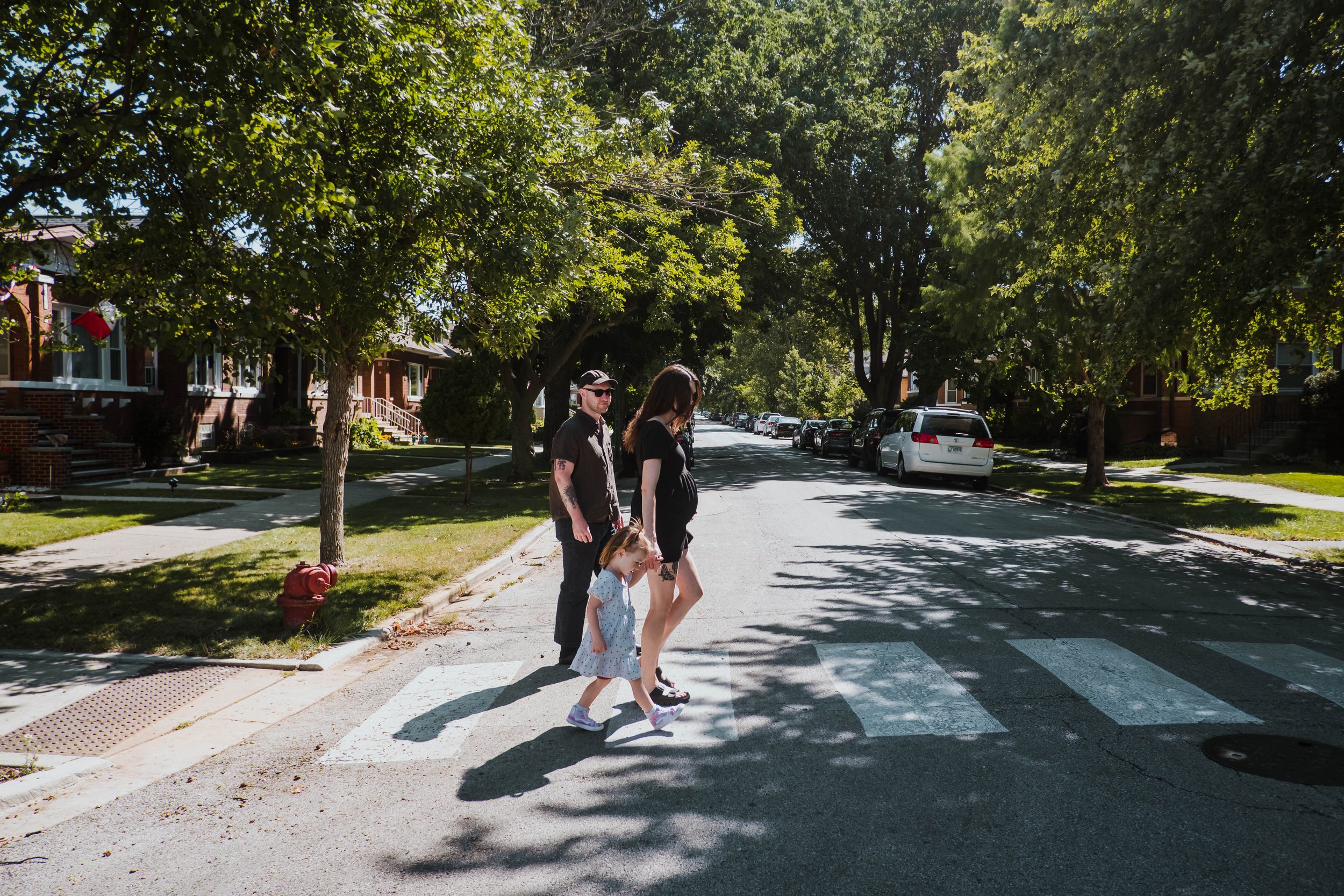 A family crossing a city street on a crosswalk on a sunny day, with trees lining the sidewalk and parked cars along the street.