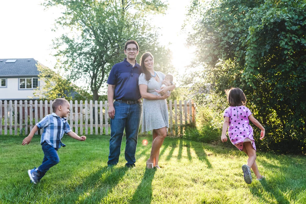 Family of five in a backyard with green grass and trees, two children running, and a woman holding a baby, with sunlight shining in the background.