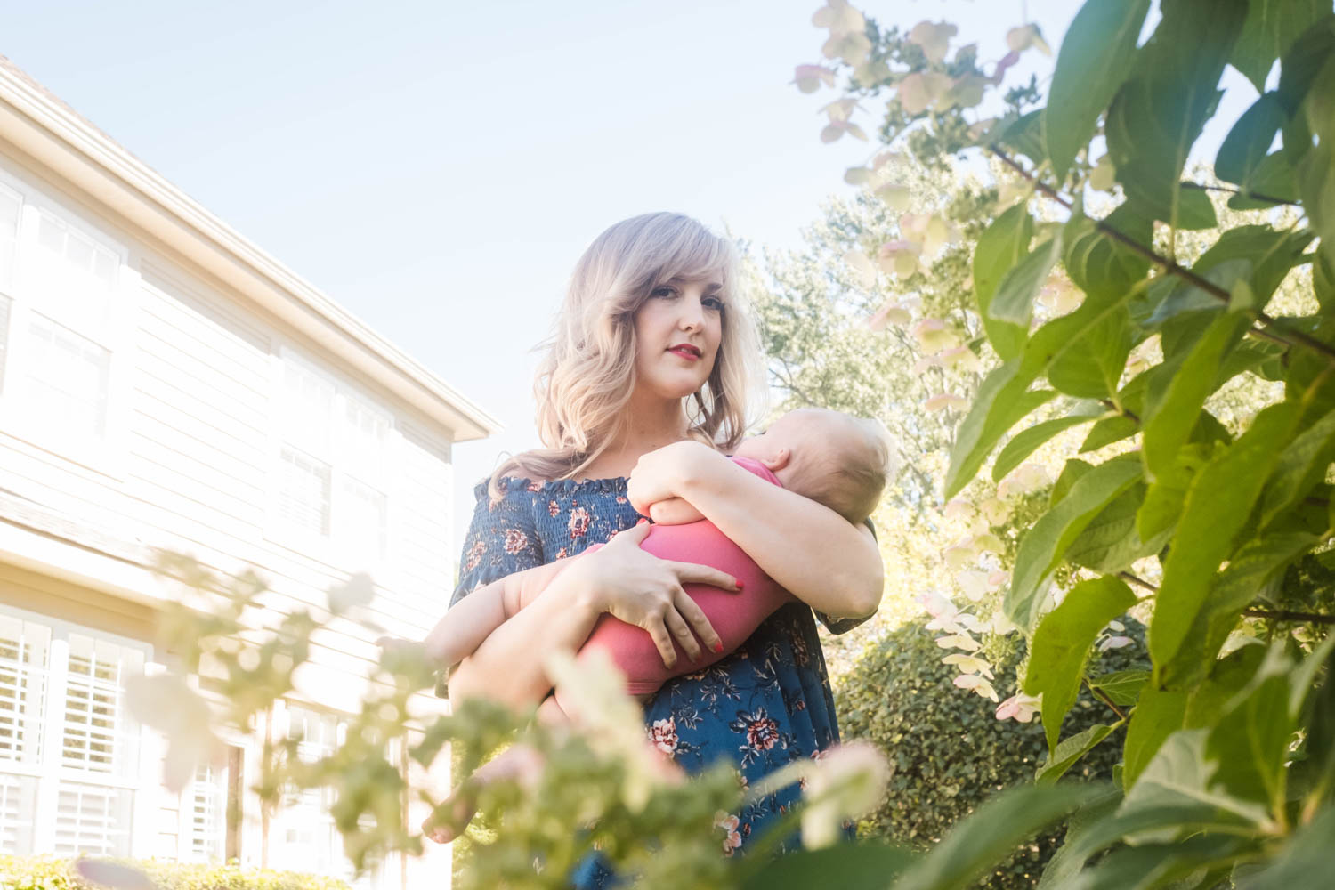 A woman with blonde hair in a blue floral dress holds a baby in a pink outfit outdoors in a garden with greenery and a large house in the background during a cinematic-family-legacy-photo-session
