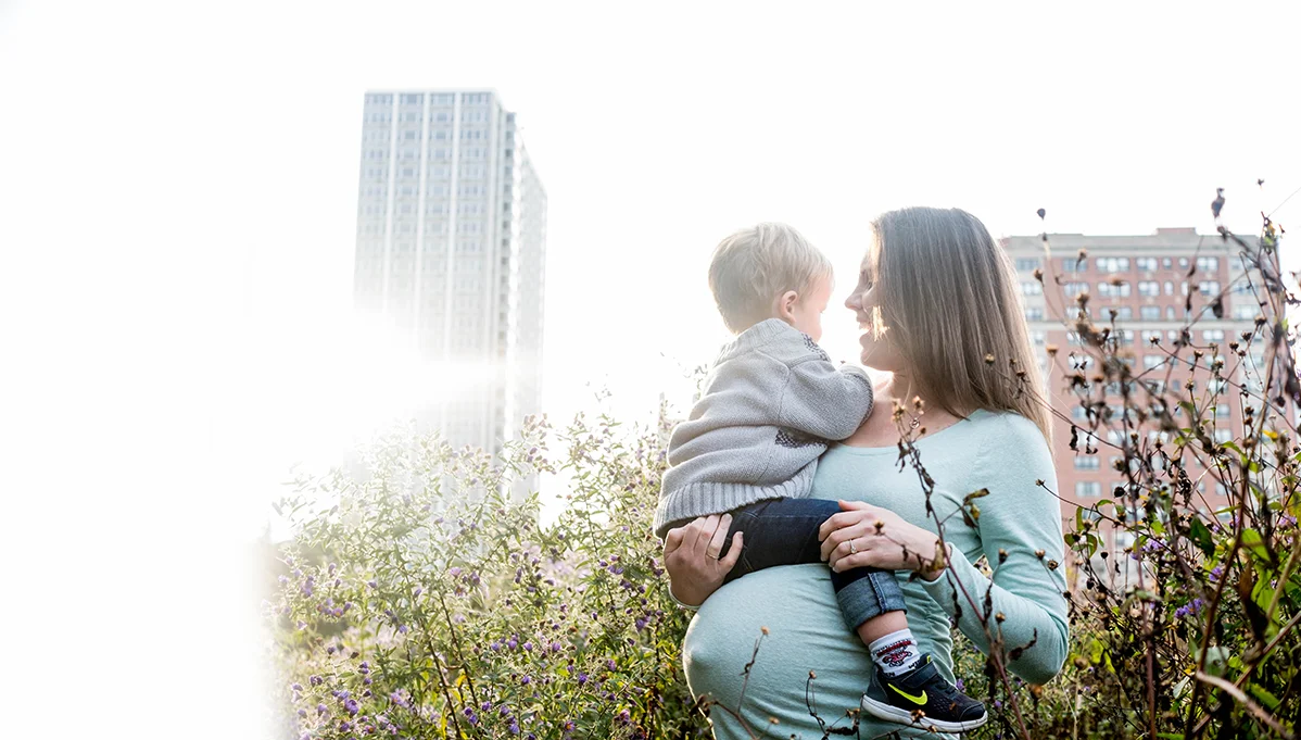 A woman holding a young child in an outdoor park area with flowering bushes, skyscrapers, and city buildings in the background, during daylight.