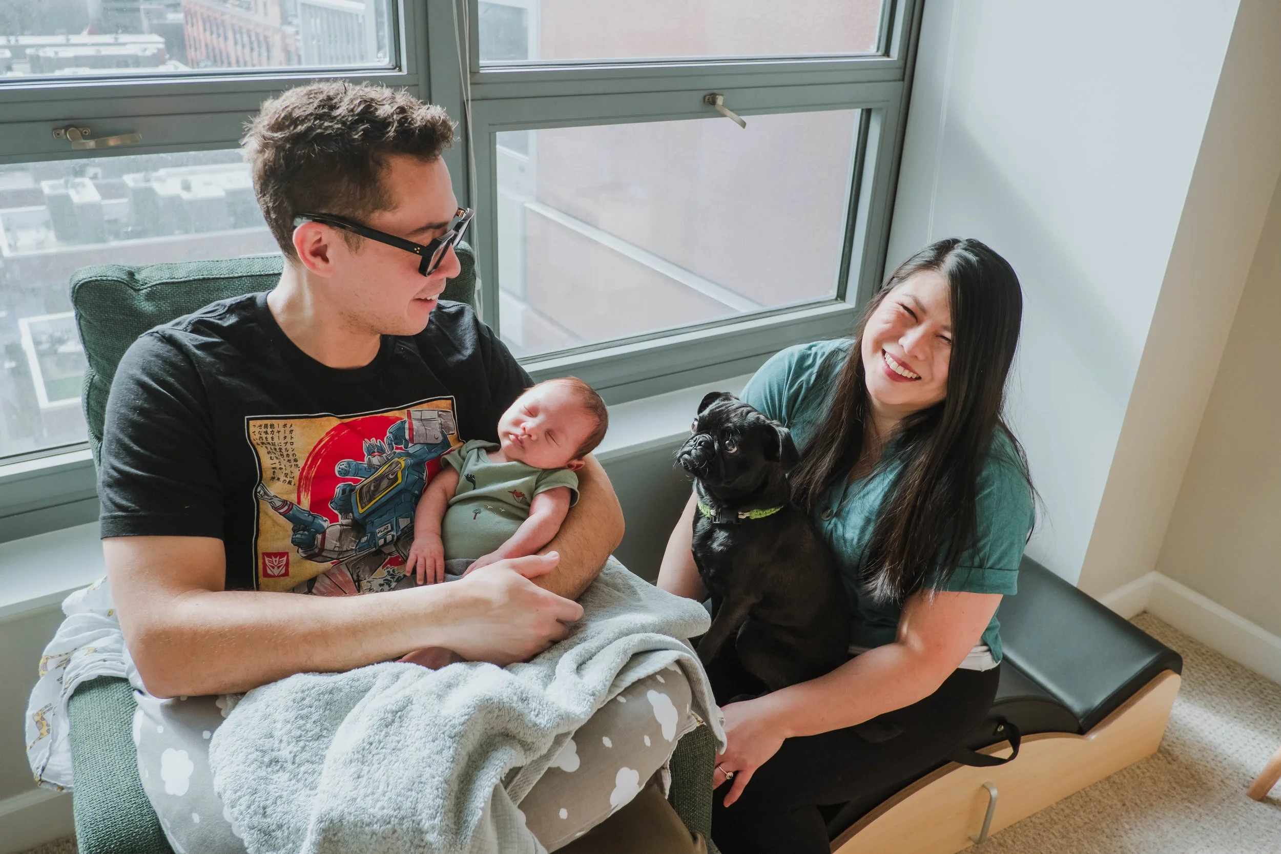 A man holding a newborn baby and a woman holding a small black dog sitting on a hospital bed near a large window in a high-rise building, with city buildings visible outside.