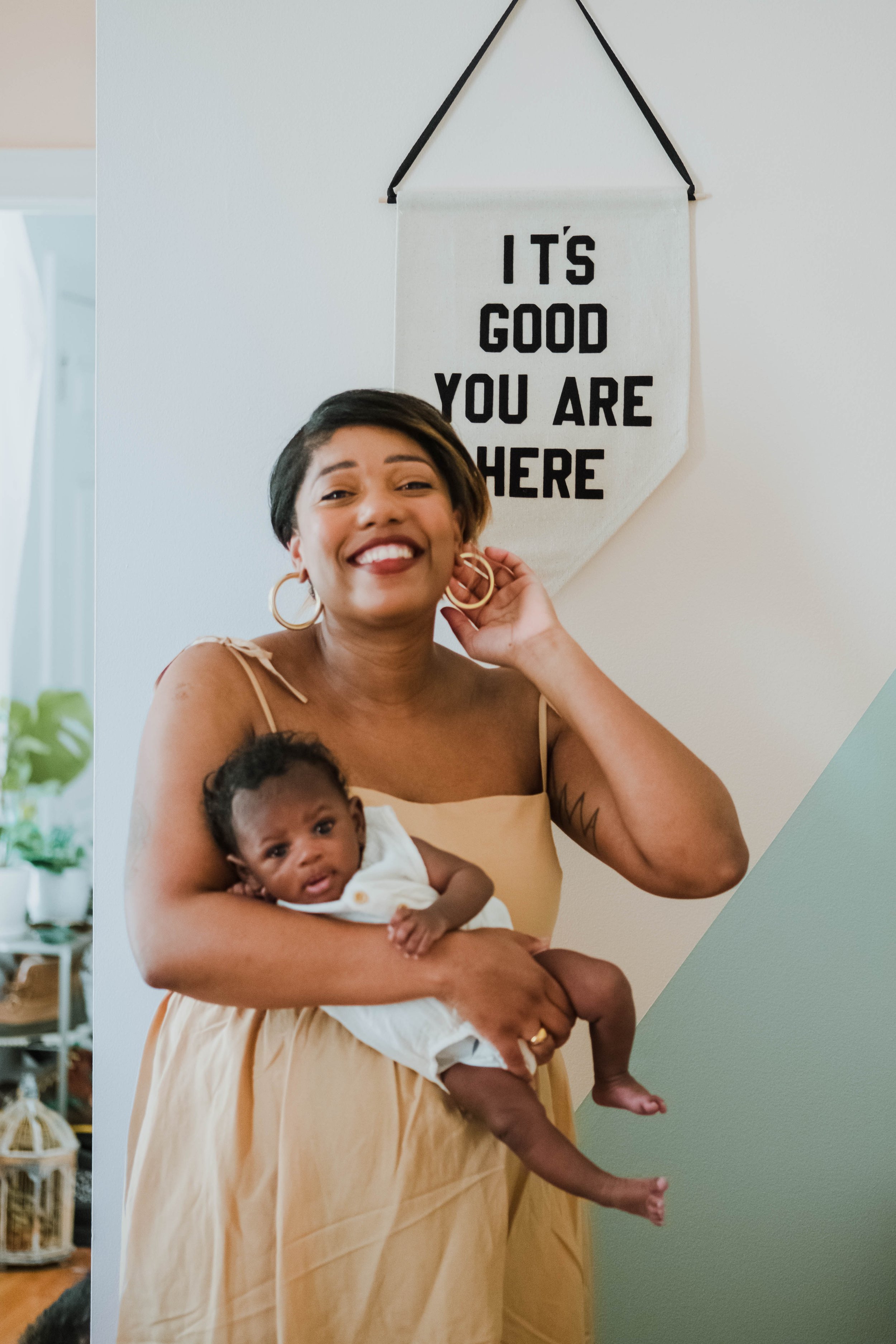A woman with short dark hair and gold hoop earrings is smiling and holding a baby girl with dark hair and dark skin, dressed in white. Behind them, a white wall features a hanging fabric banner with the text 'IT'S GOOD YOU ARE HERE' in black.