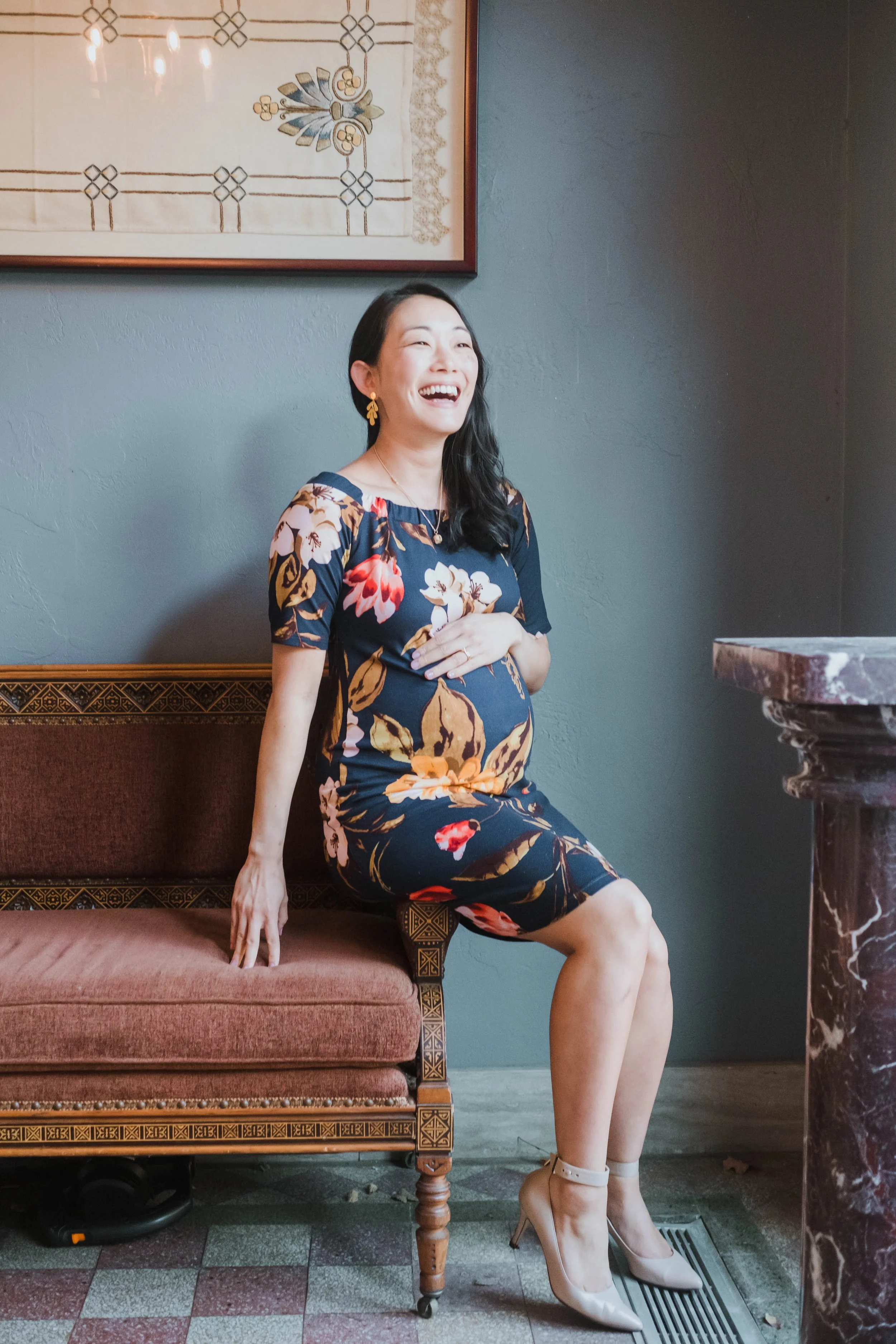 A woman in a floral dress sitting on a vintage style pink and gold bench, laughing, with her hand on her stomach. She is wearing high heels and earrings.