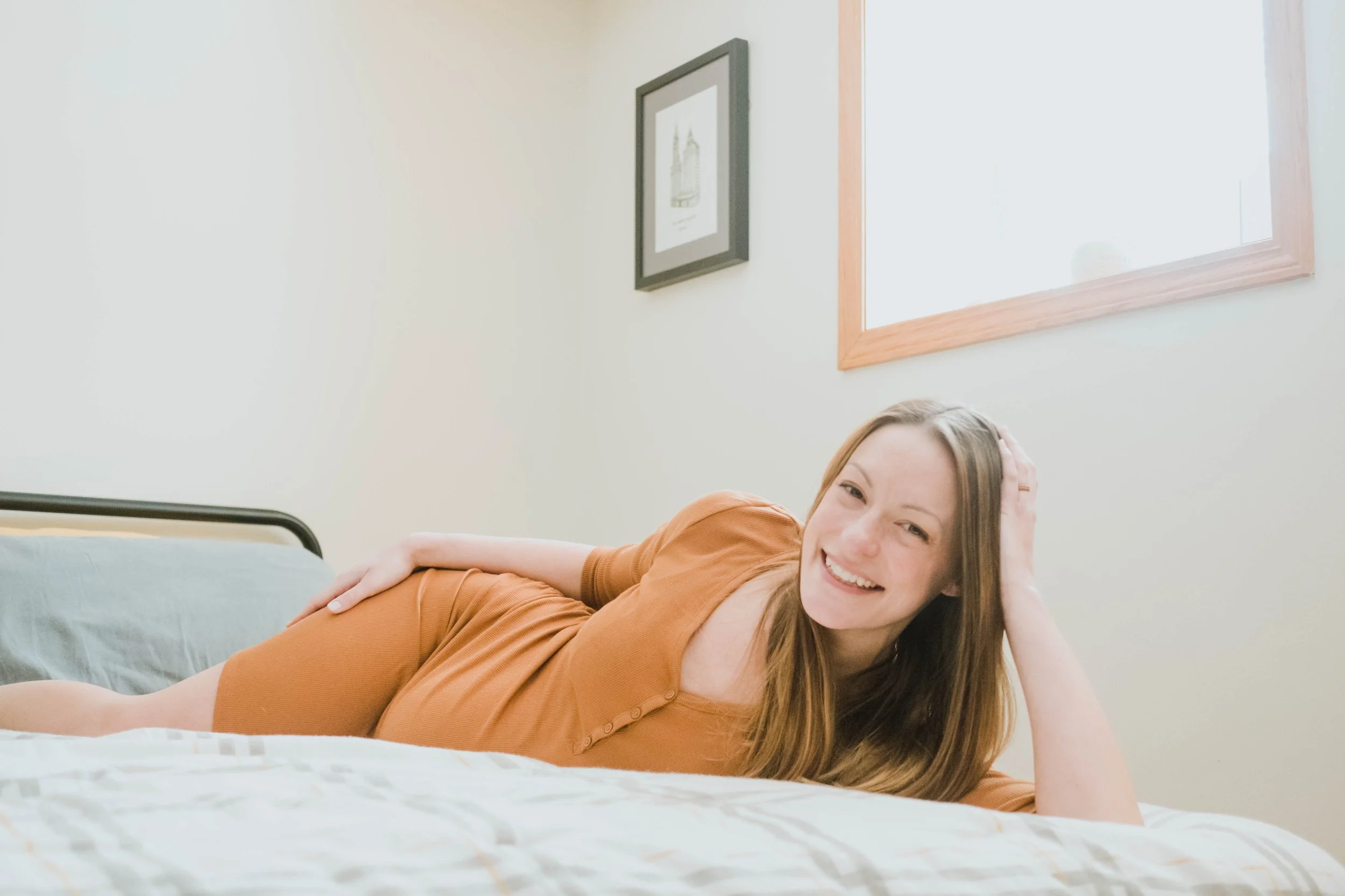 A young woman with long red hair lying on her side on a bed, smiling and resting her head on her hand. She is wearing an orange top and is in a room with a window and framed pictures on the wall.