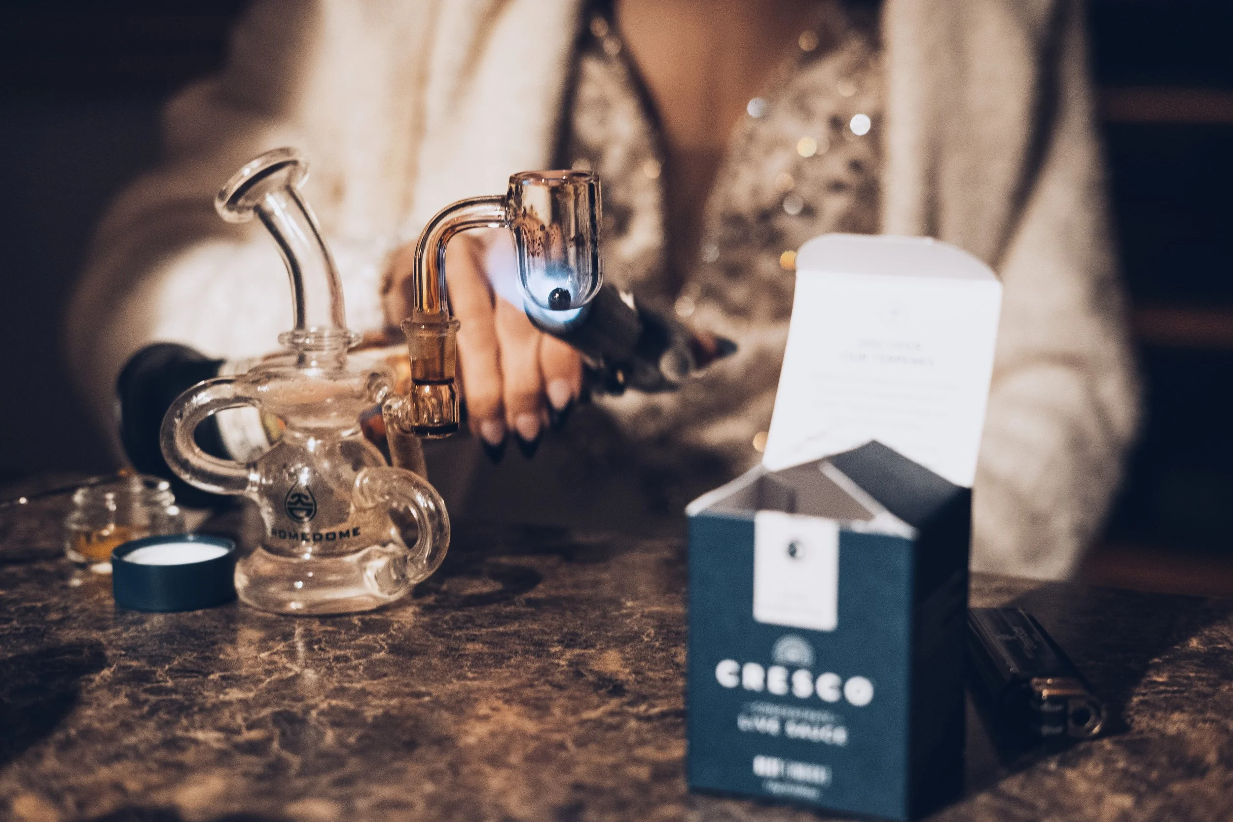 Close-up of a person holding a glass bong, with a small box of Cresco Live Resin Carts on a dark wooden table. The person's hand is near the bong, and the background is blurred.