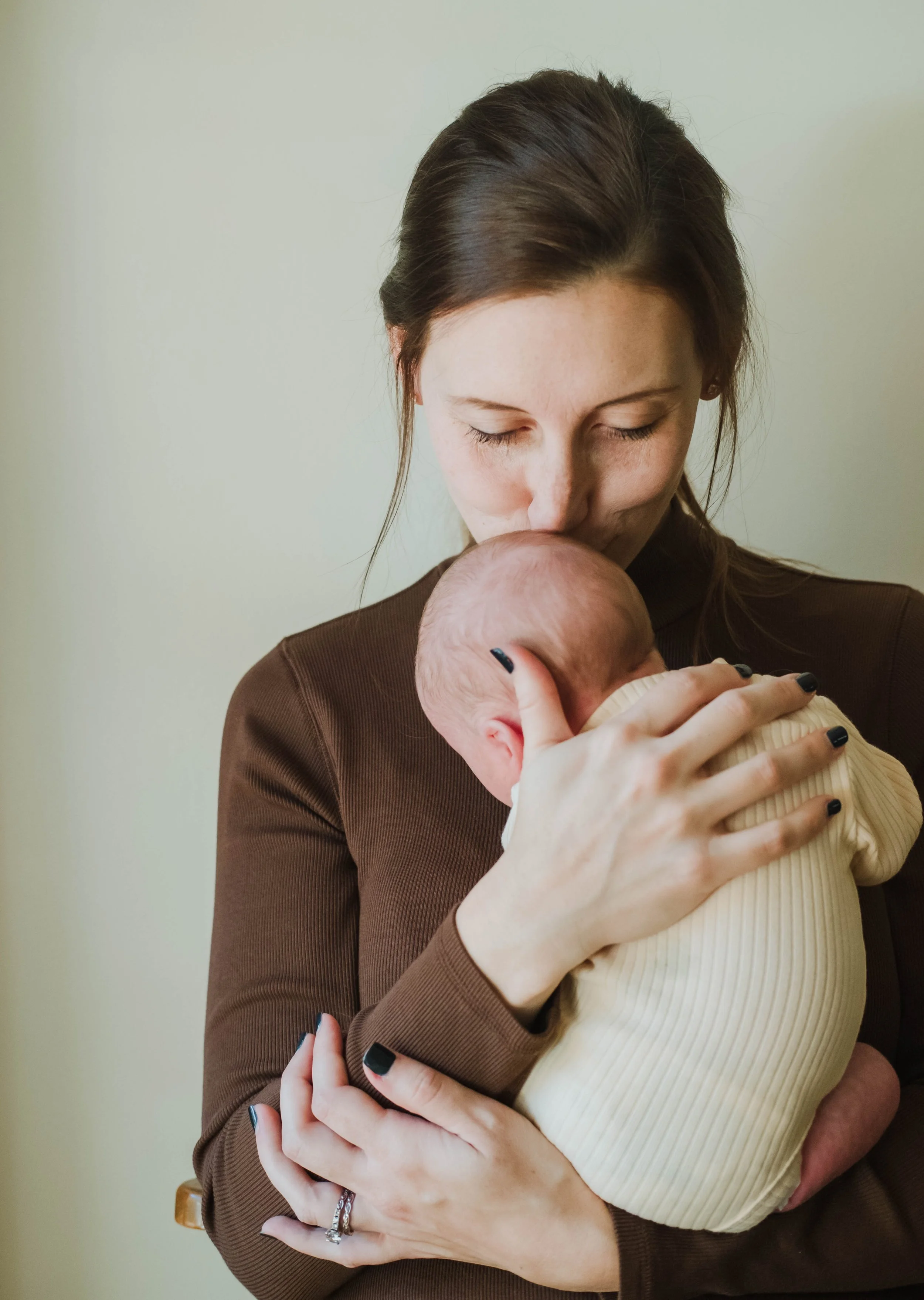 A woman with dark brown hair in a bun lovingly holding a bald baby close to her face, her eyes closed in an affectionate pose.