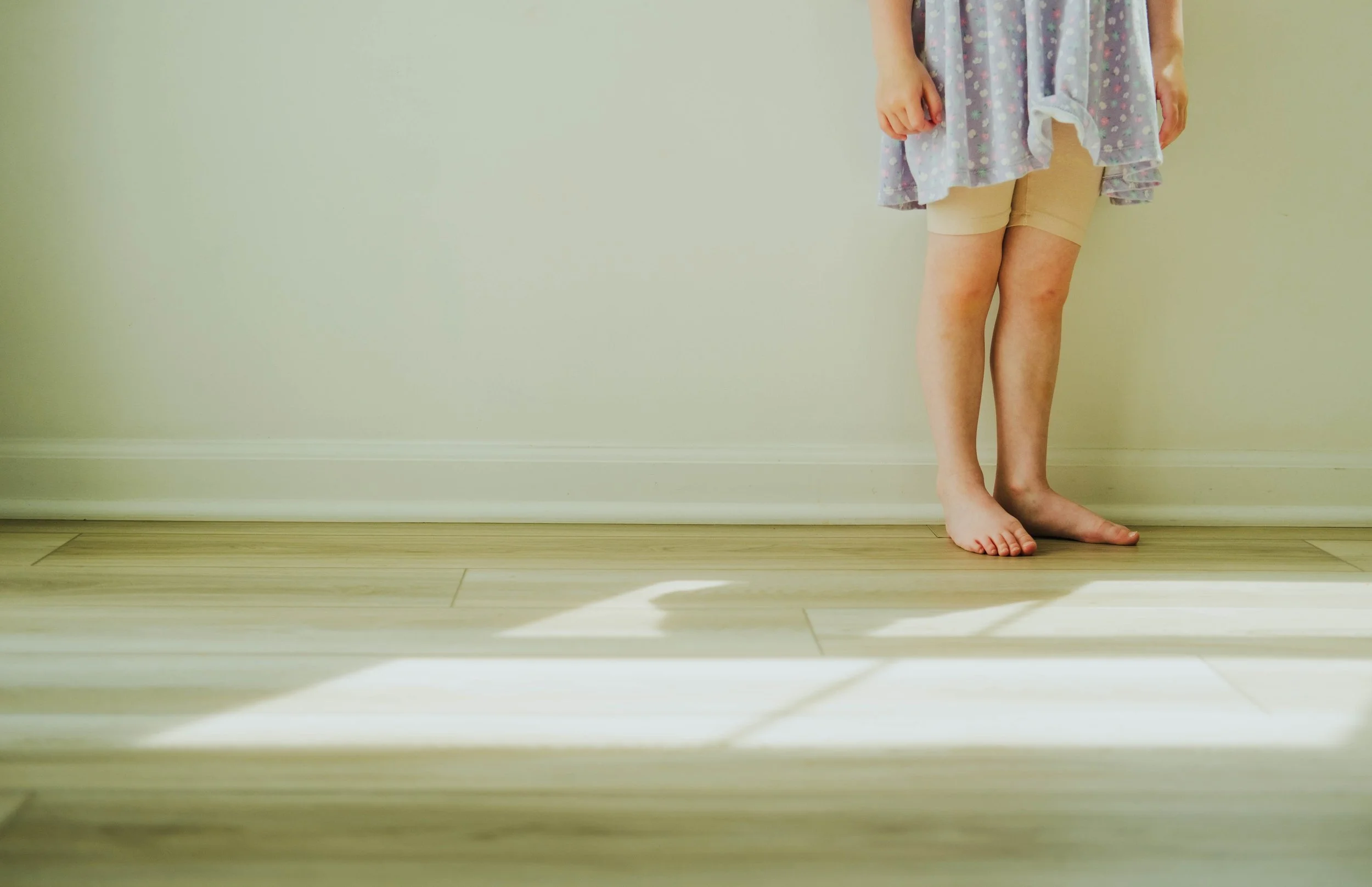 A child's lower body, barefoot, standing on a light wood floor next to a plain cream-colored wall, wearing tan shorts and a pajama top with a dark background and white and purple polka dots.