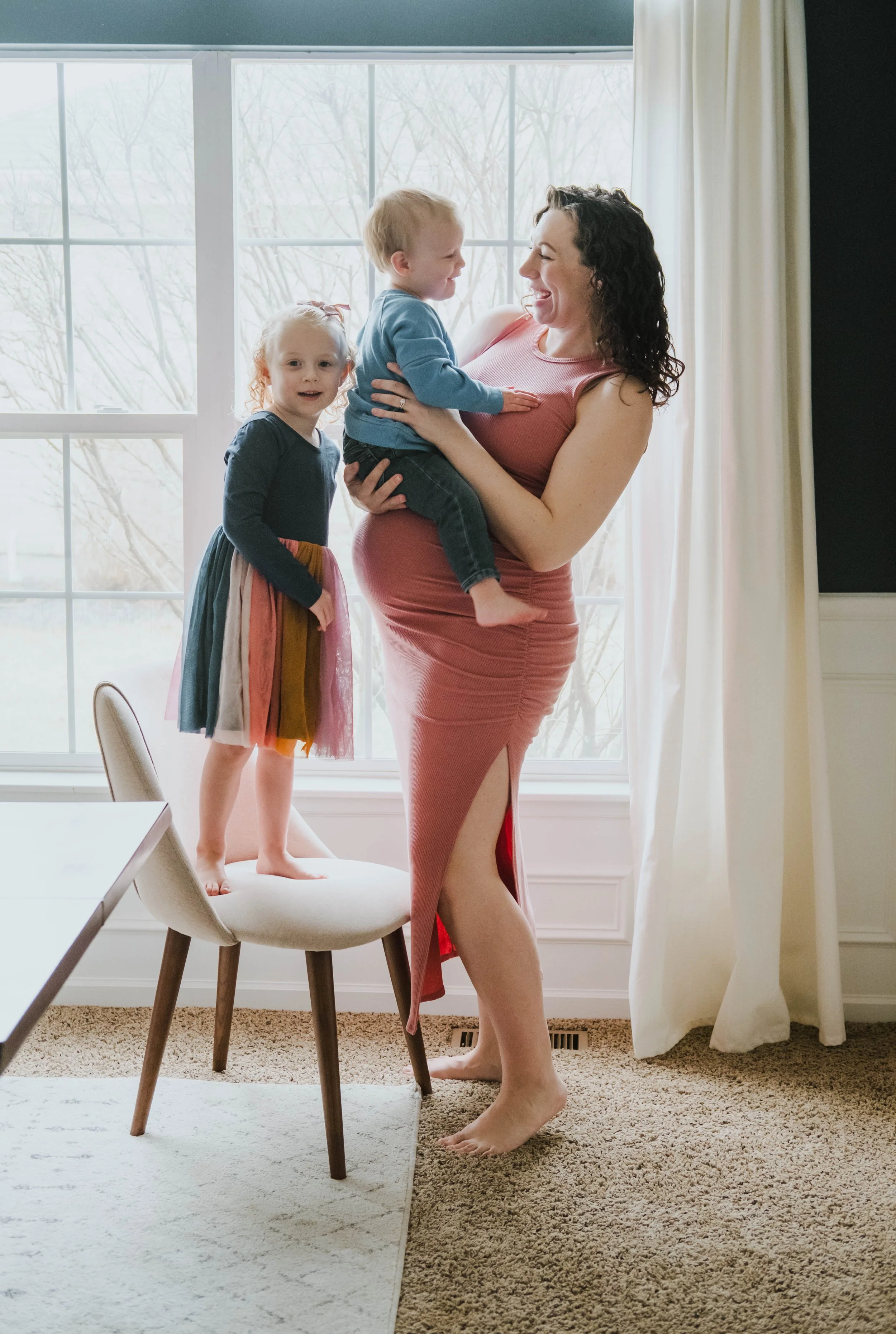 A woman holding a young boy and standing beside a girl who is standing on a chair by a large window with curtains; the woman and children are smiling and engaging with each other in a bright room.