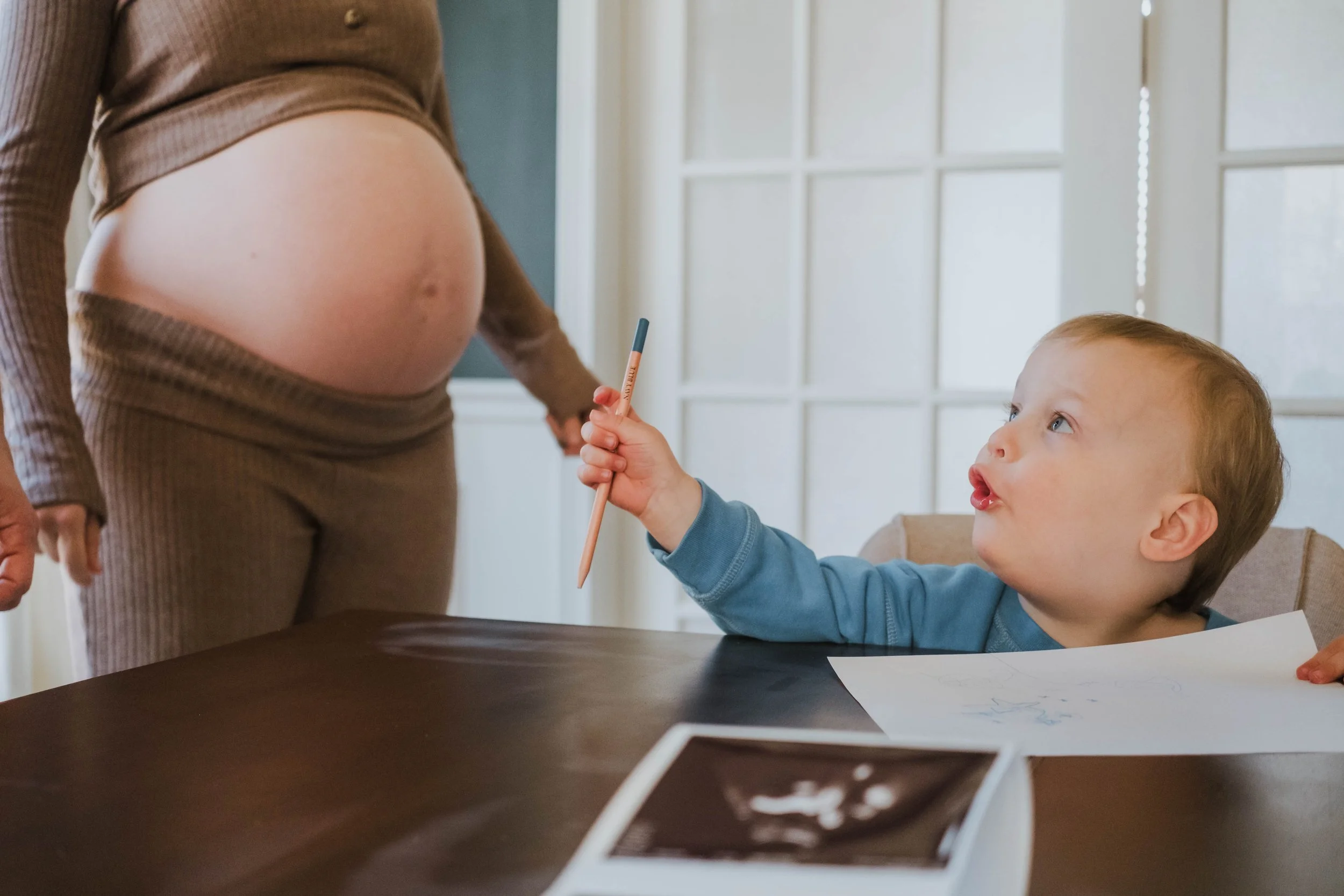 A pregnant woman stands near a young child sitting at a table. The child is holding a pencil and appears to be drawing or writing on paper, looking up at the woman with curiosity.