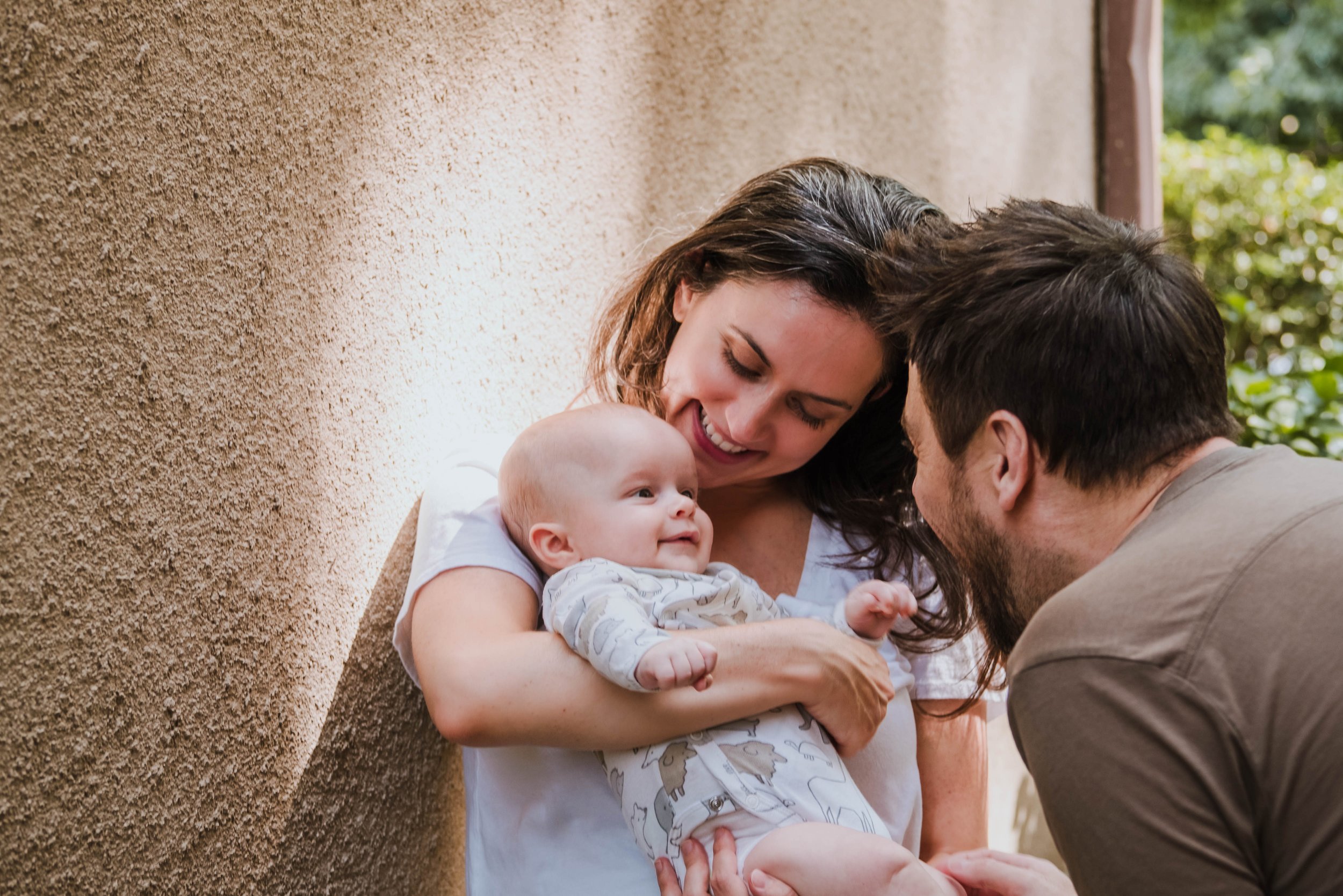 A family with a woman, man, and baby engaging in a tender moment outdoors, smiling and looking at each other.