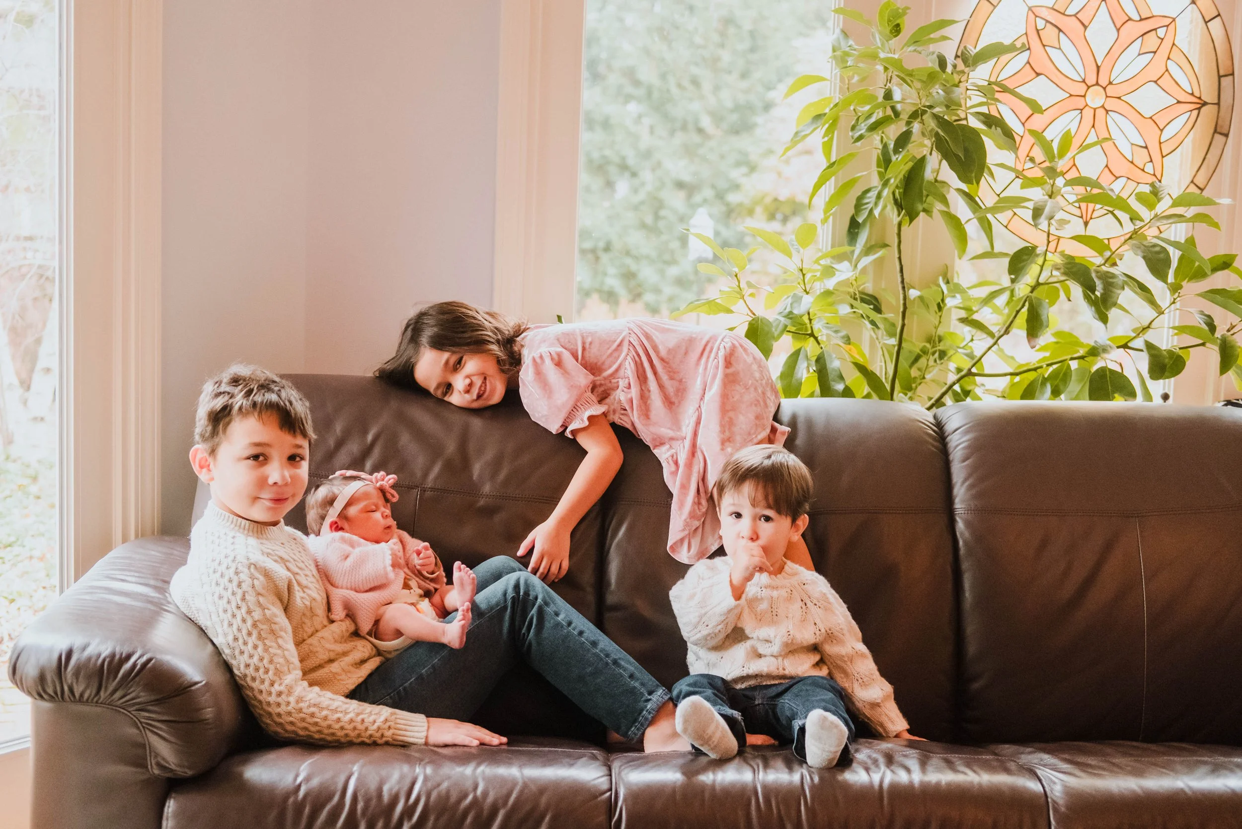 A woman and four children sitting on a brown leather couch in a living room with large windows and a decorative wall hanging, surrounded by green indoor plants during a cinematic-family-legacy-photo-session
