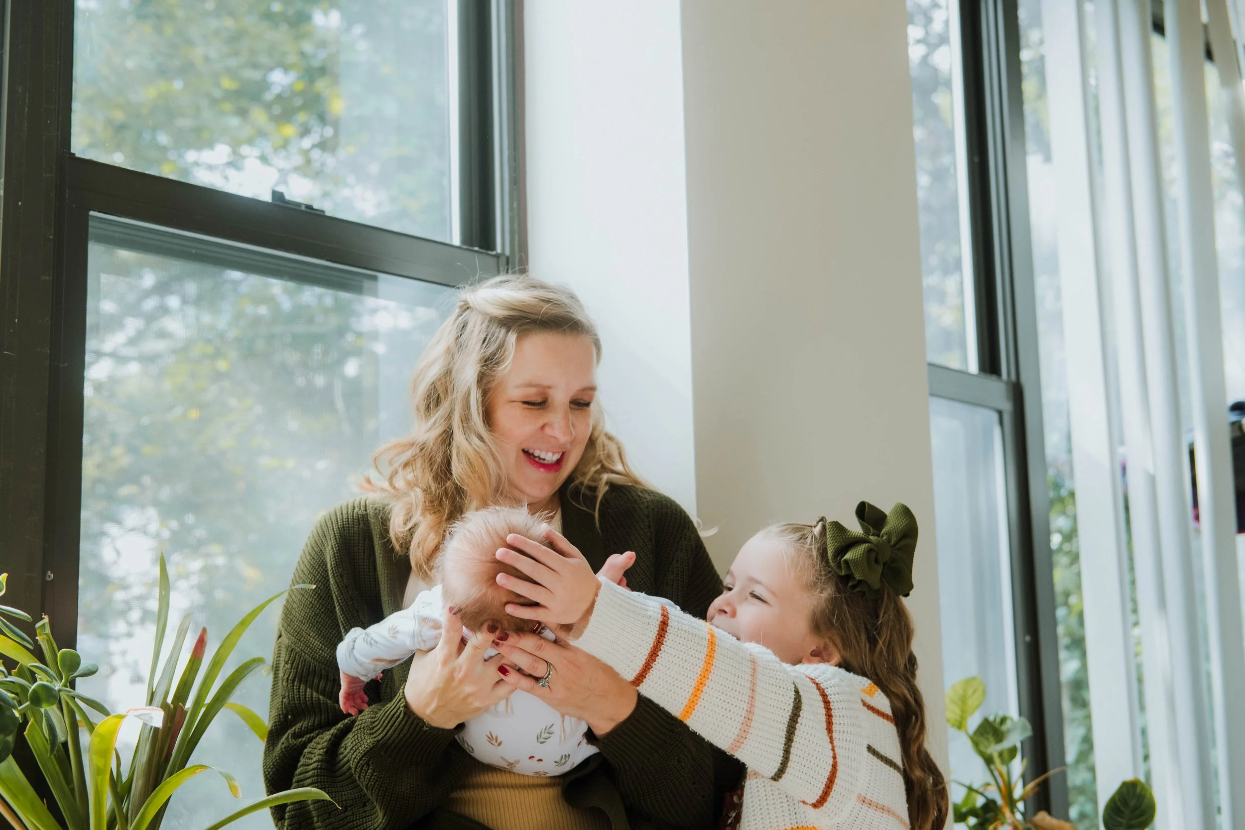 A woman with curly blonde hair smiling as she holds a small child's head. The child is reaching out with both arms. Another girl with long hair and a large green bow in her hair is also reaching towards the child. They are indoors near large windows 