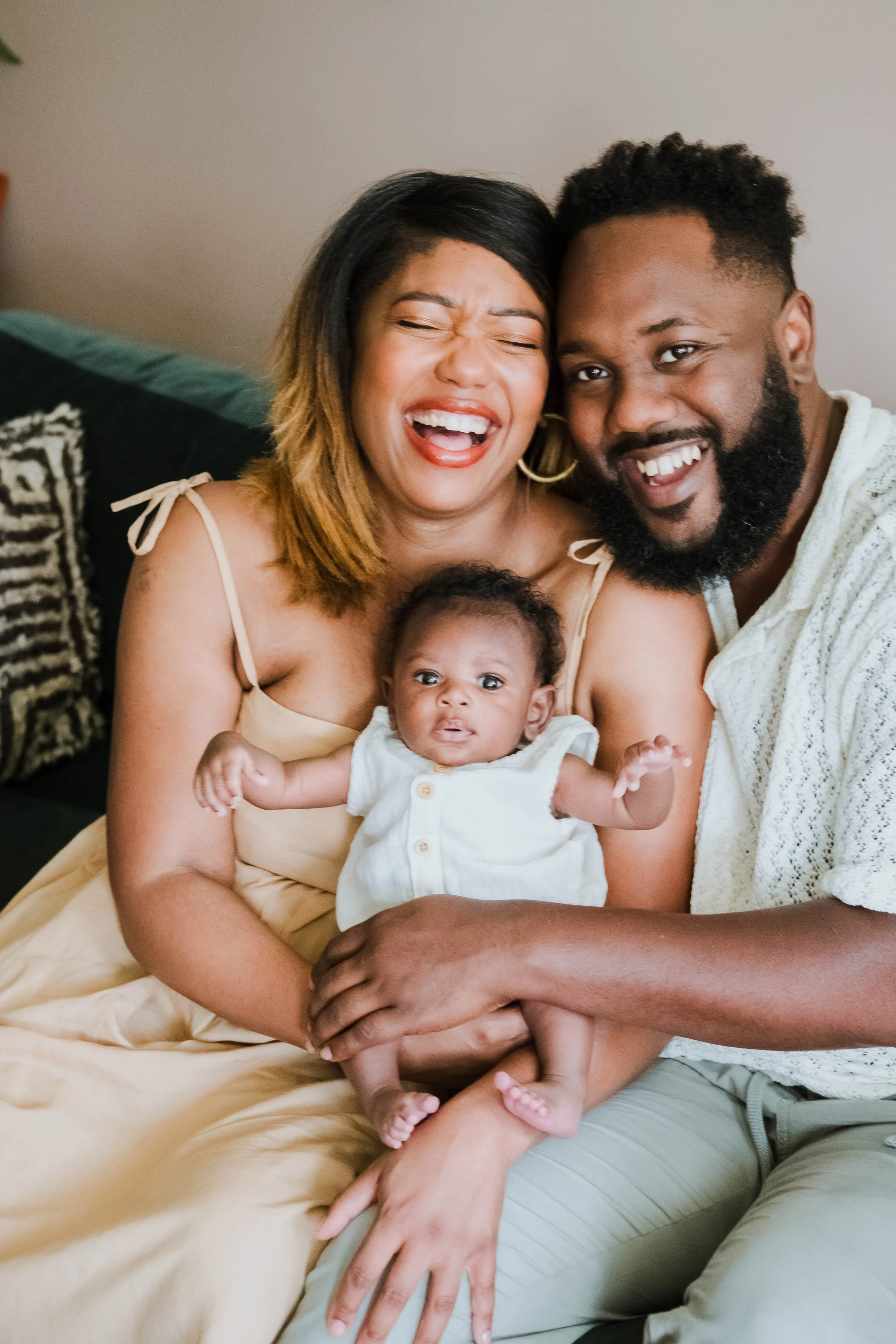 A smiling couple with a baby, all sitting close together on a couch in a cozy home setting.