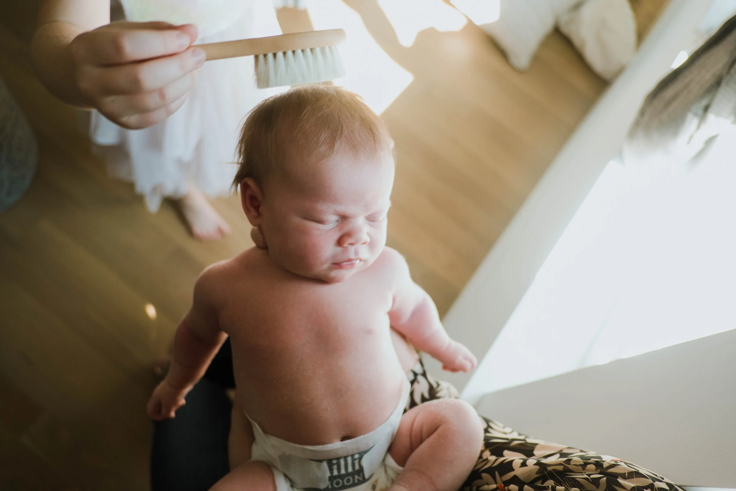 A small, shirtless baby with red hair sitting on a person's lap, eyes closed, while someone brushes the baby's hair with a wooden hairbrush in a brightly lit room with wooden flooring.