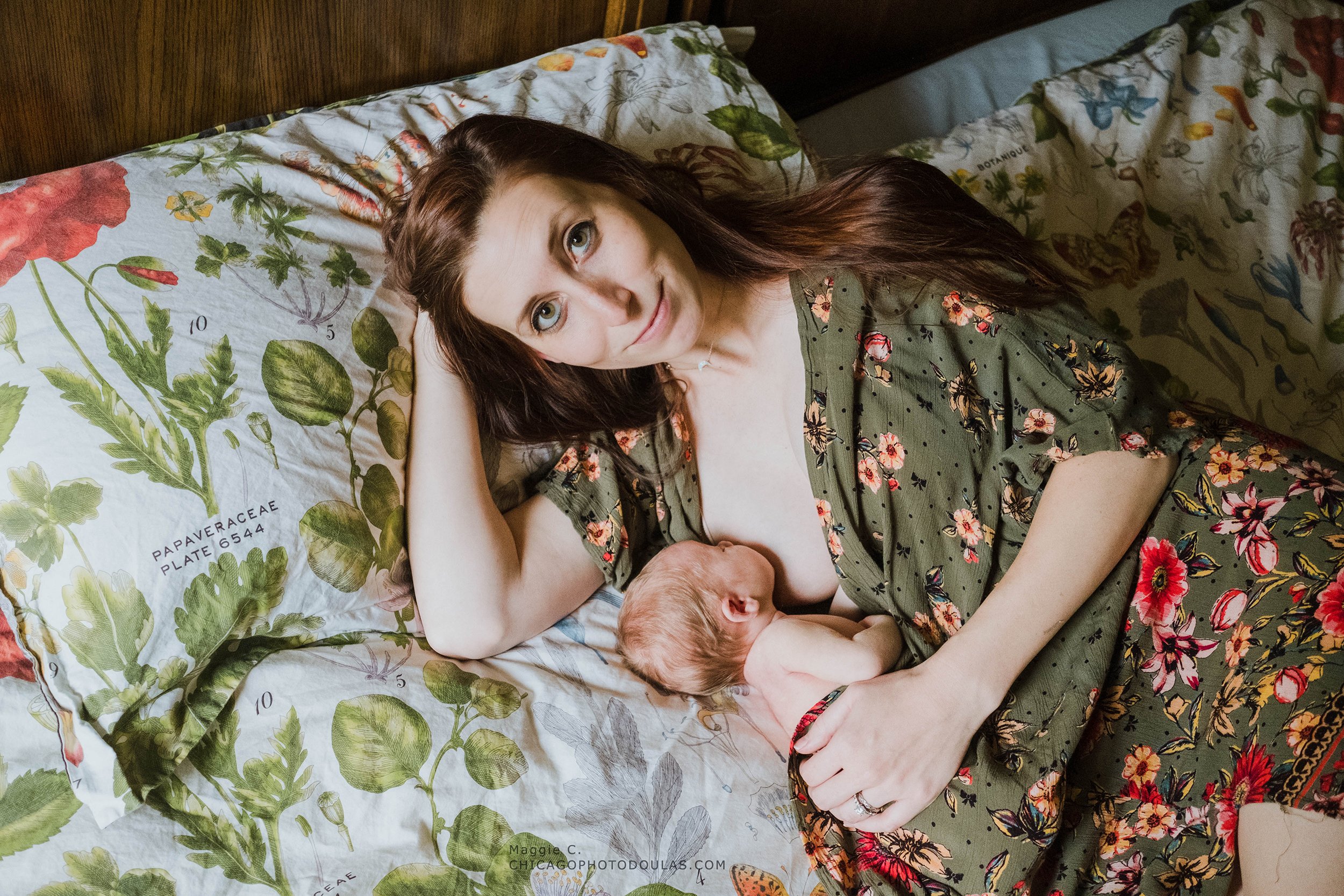 A woman with long brown hair and blue eyes lying on a bed, resting her head on her hand, looking at the camera. She is wearing a green dress with pink floral patterns. Beside her is a sleeping baby with red hair, lying close to her.