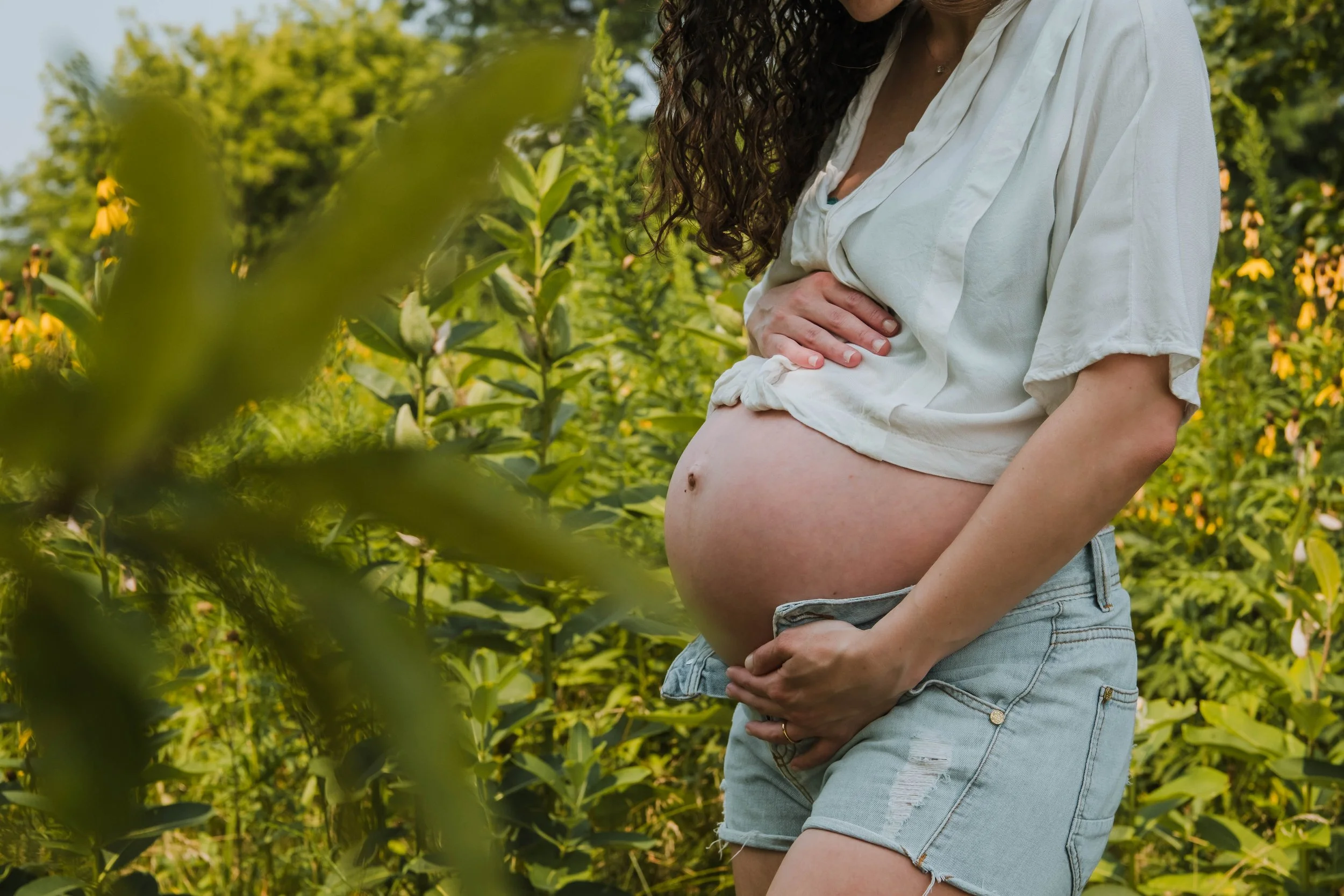 A pregnant woman with curly hair wearing a white blouse and denim shorts standing in a lush green garden.