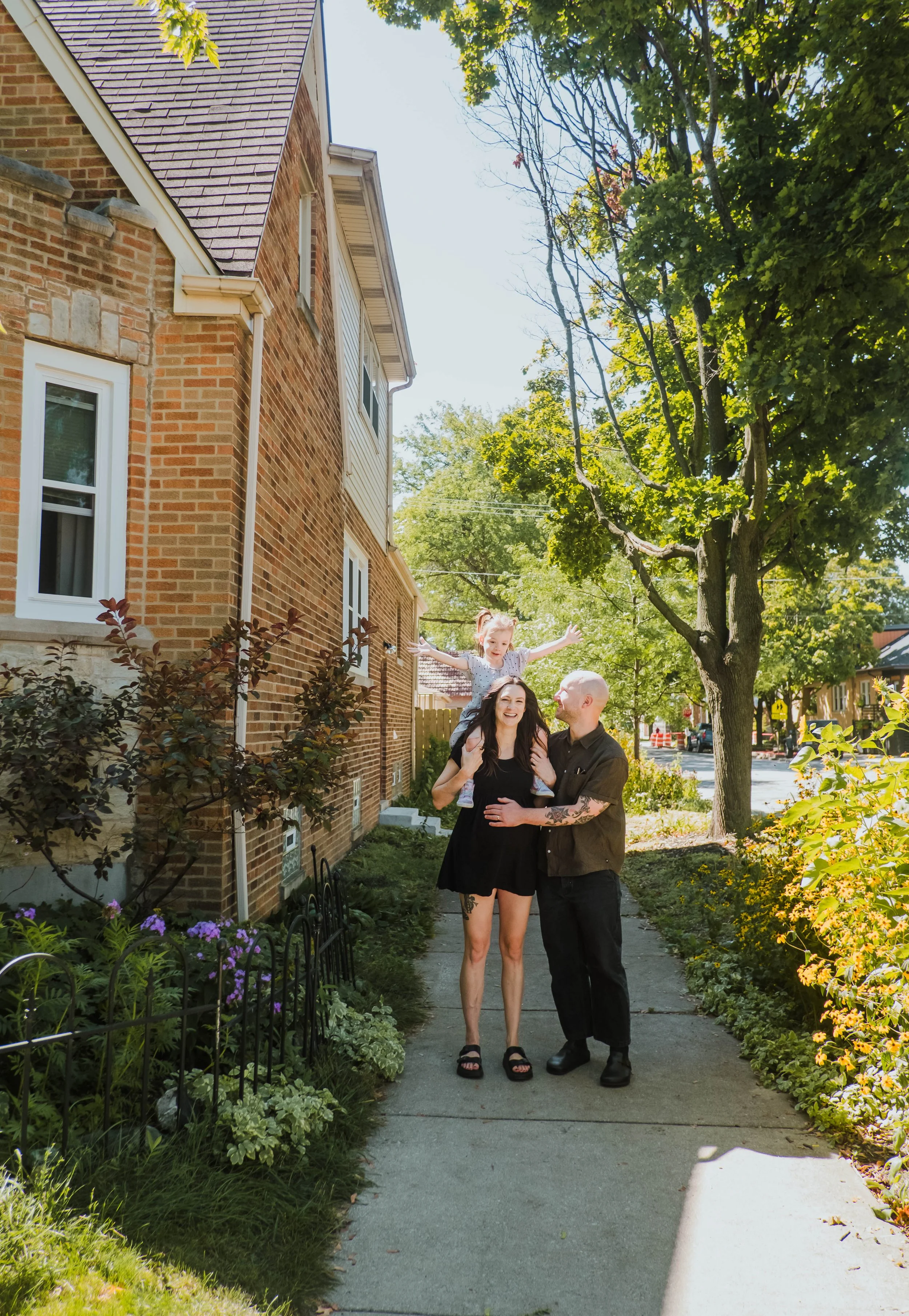 A family of three, consisting of a woman, a man, and a young girl sitting on the woman's shoulders, standing on a sidewalk during daytime. They are smiling and enjoying a sunny day near a red brick house with windows. There are trees and flowering pl