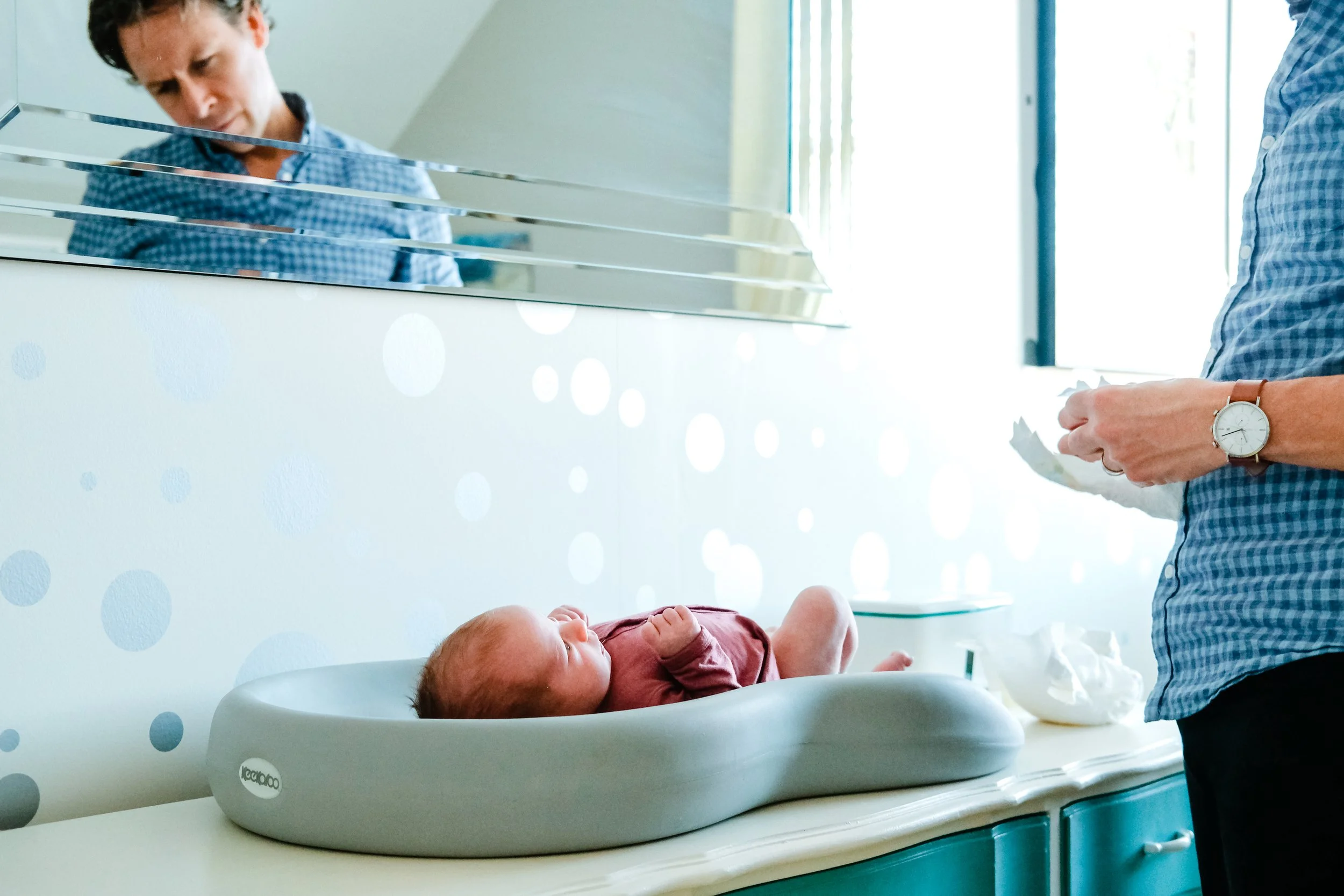 A baby lying in a white changing table while a woman, whose face is partially visible, stands nearby holding a tissue or cloth. The woman is wearing a blue checkered shirt and a watch on her left wrist. The room has bright natural light coming throug