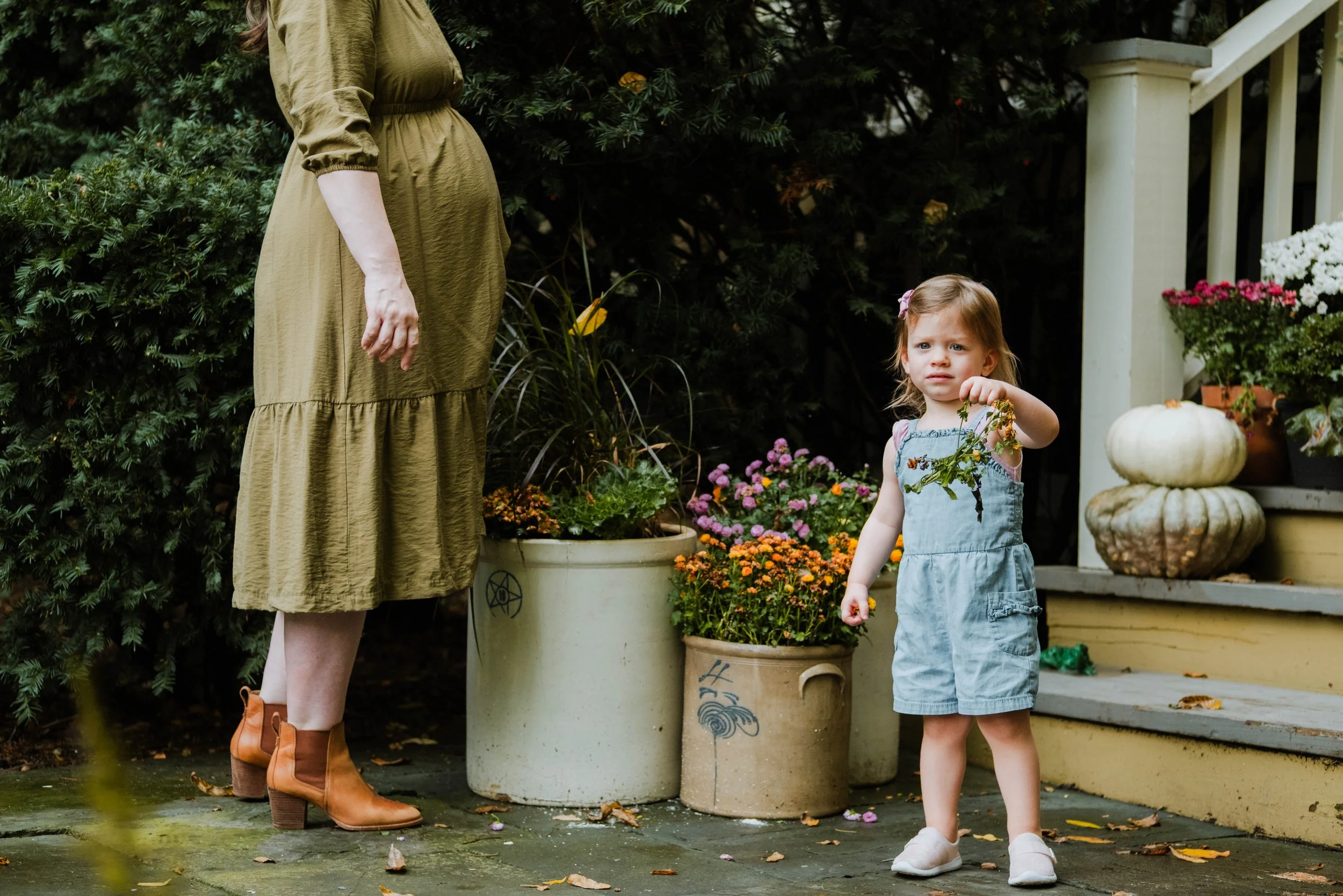 A young girl with light brown hair, wearing a light blue dress and white shoes, standing outdoors near potted plants and pumpkins, holding a small plant with orange flowers. An adult woman wearing a long olive green dress and brown boots is partially
