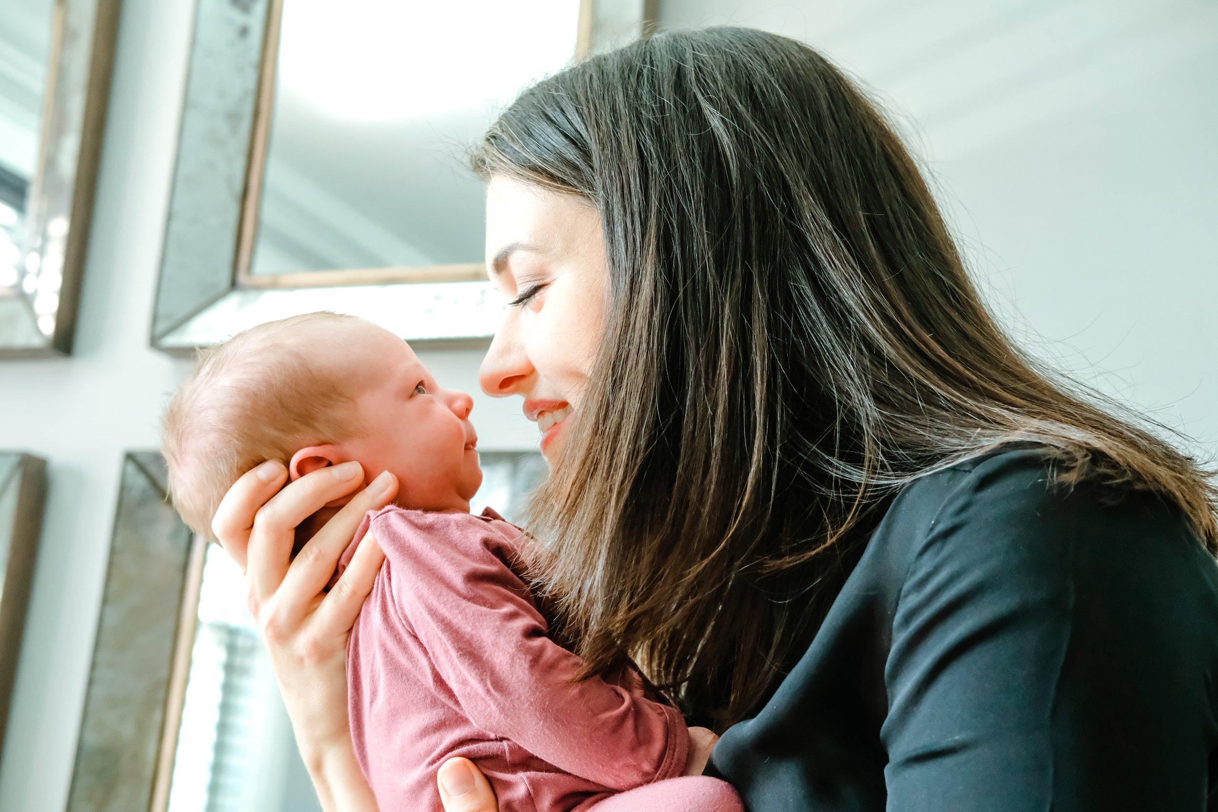 A woman with long dark hair holding an infant close to her face, smiling and looking at each other in a bright room with windows and mirrors.
