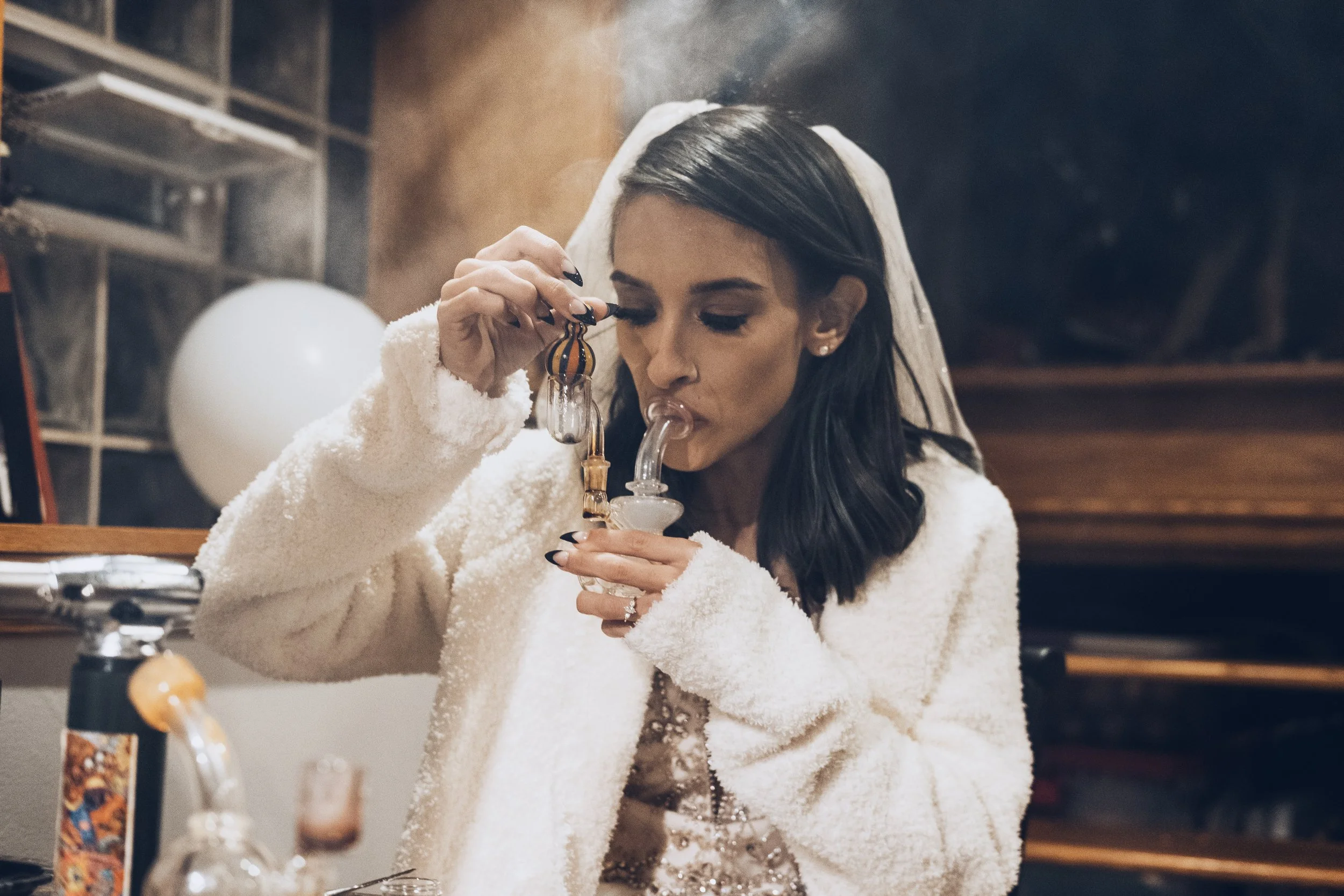 Woman with dark hair wearing a white fuzzy coat and veil, using a glass bong in a kitchen with bottles and kitchen shelves in the background.