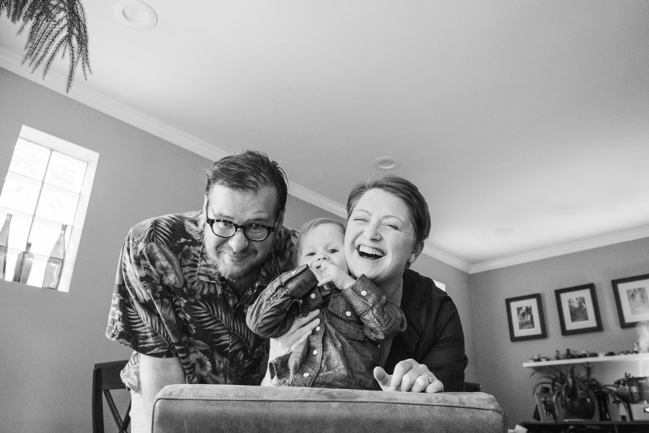 A black-and-white photo of a family, including a man with glasses, a woman, and two children, in a cozy living room, smiling and kissing.