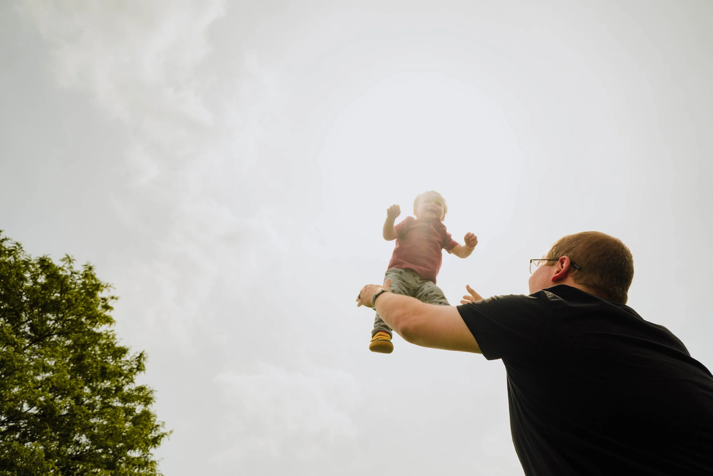 A man holding a young child in the air outdoors beneath a cloudy sky, with trees visible on the left side.