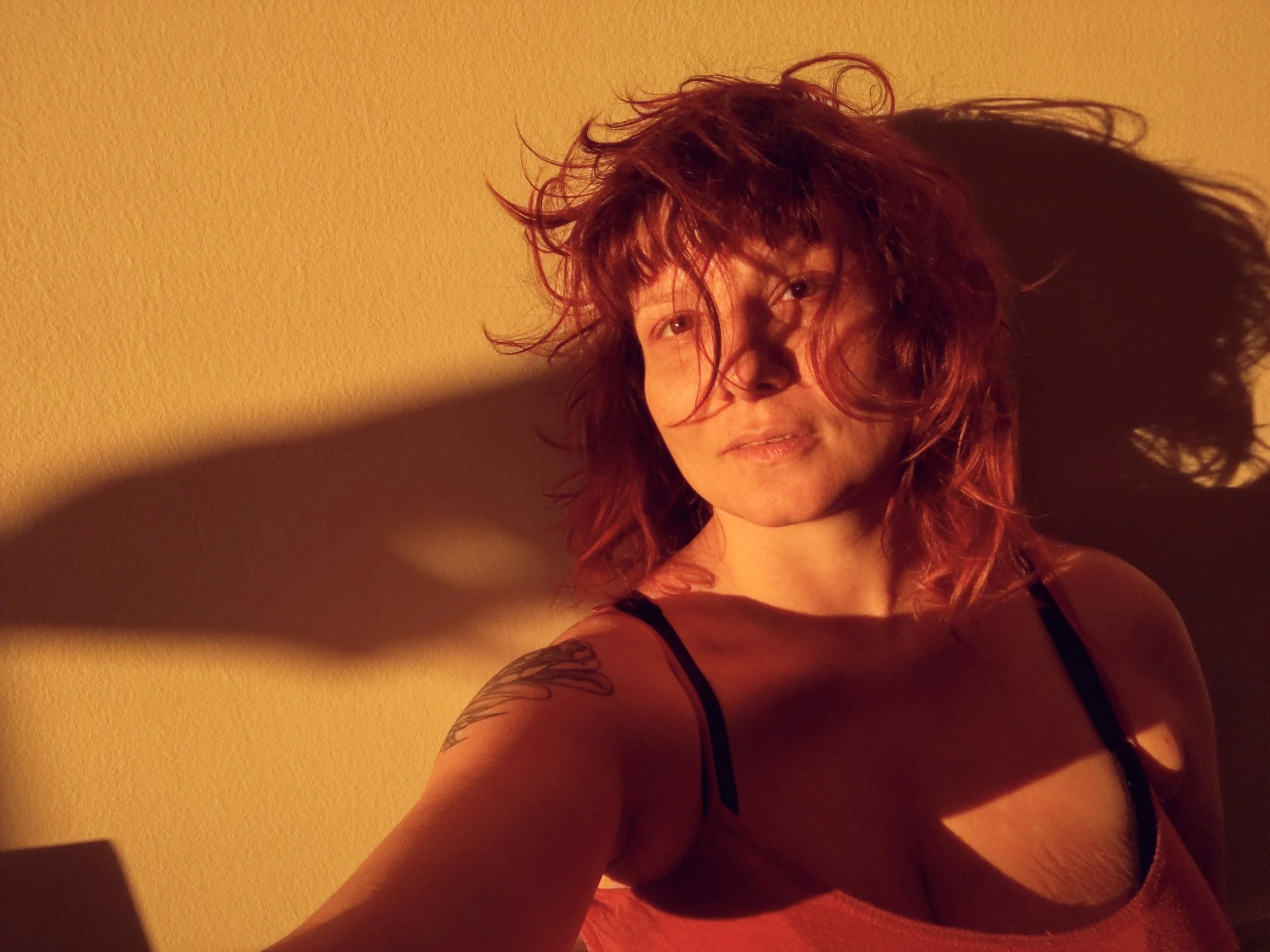 A woman with curly red hair and tattoos on her arm posing against a yellow wall, wearing a black tank top.