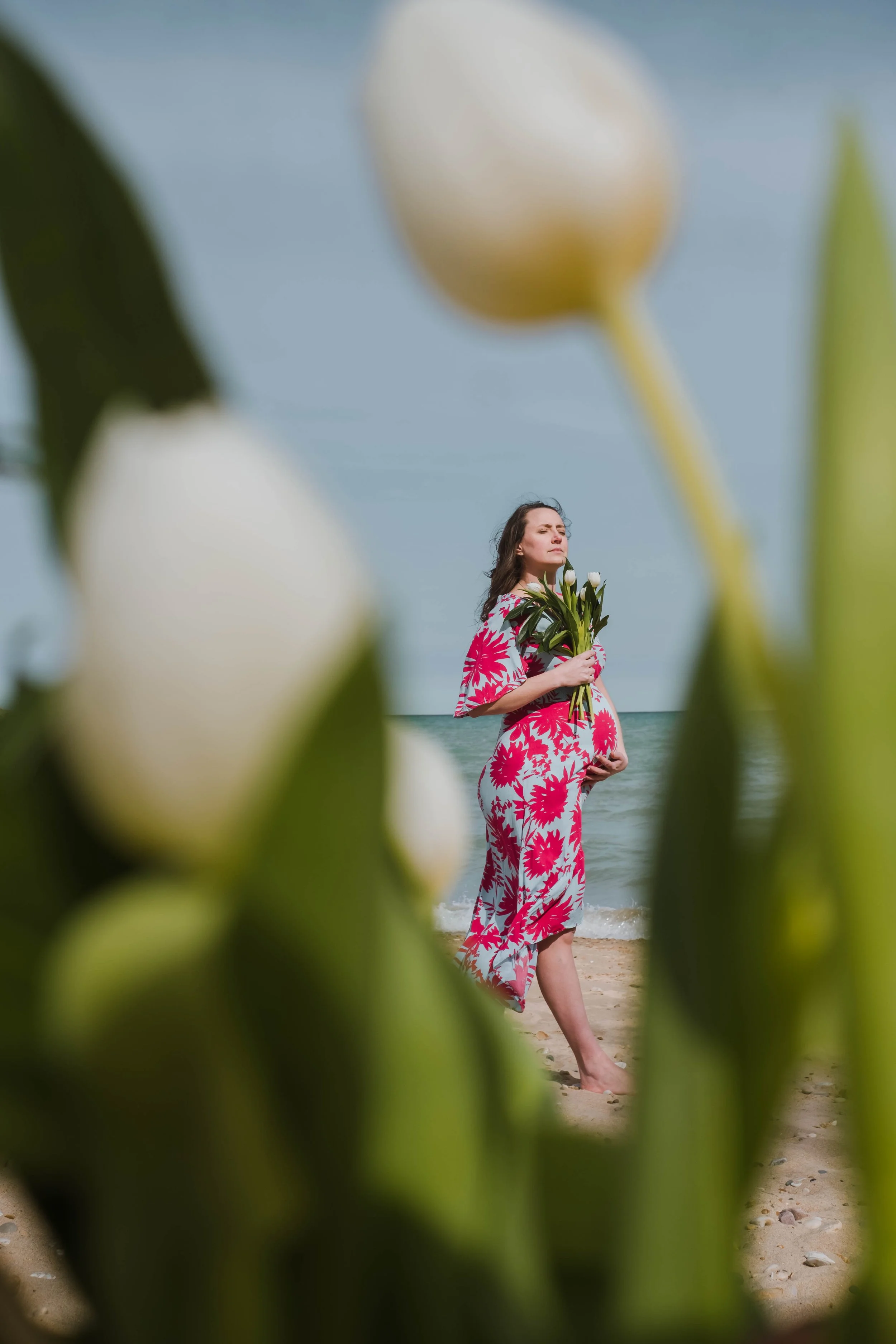 A woman in a pink and white floral dress standing on the beach, holding a bouquet of white flowers, with the ocean and sky in the background, framed by blurred green leaves in the foreground.
