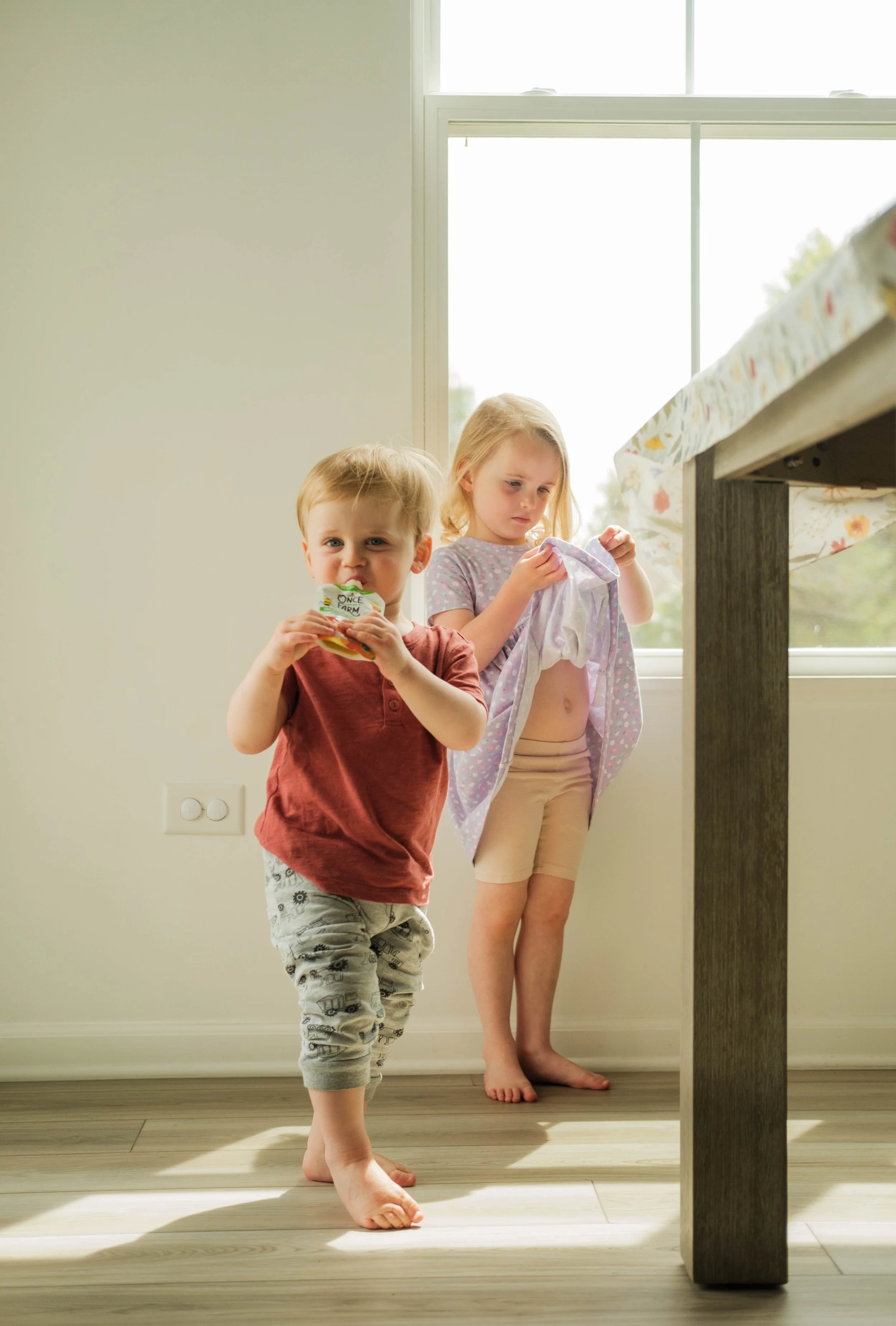 Two young children, a boy and a girl, are in a bright room near a window. The boy is holding a snack and looks at the camera, while the girl, barefoot, is examining a piece of clothing she is holding.