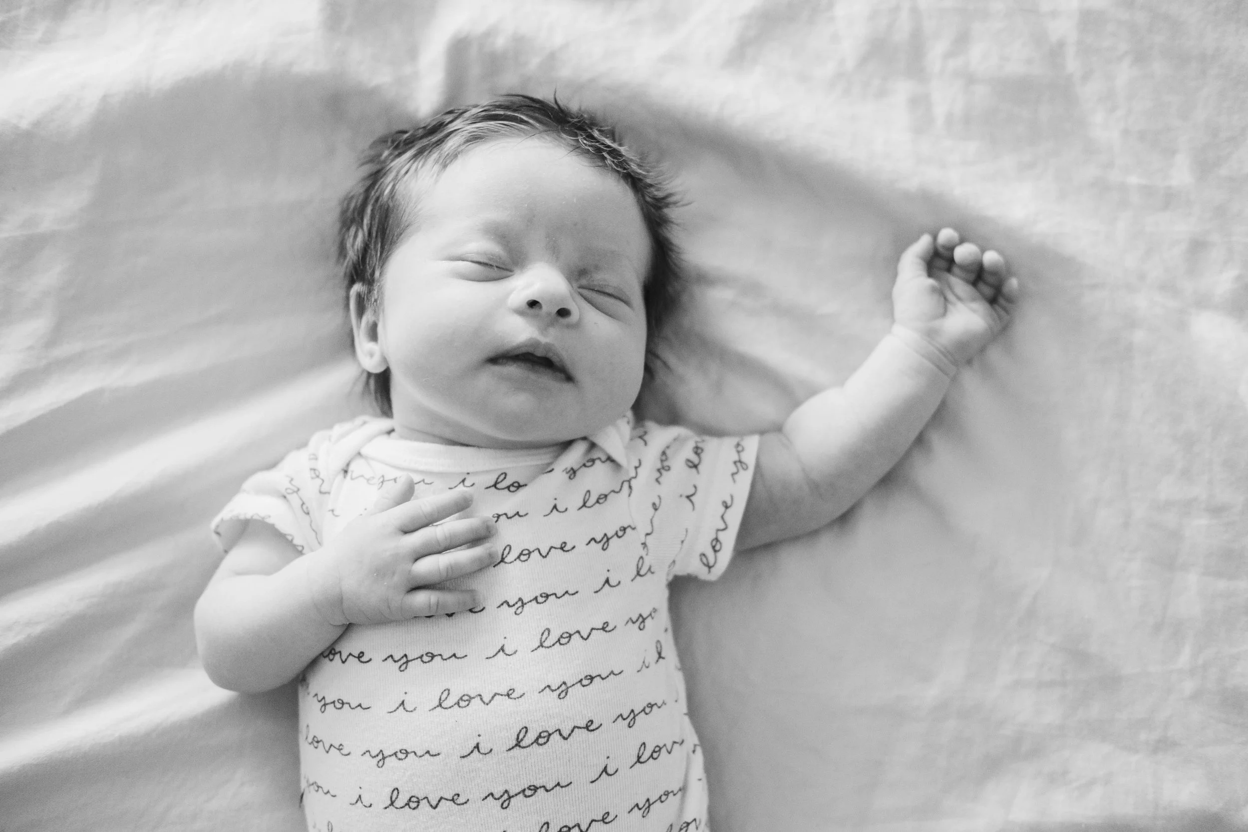A black-and-white photo of a sleeping newborn baby with closed eyes, lying on a soft surface, wearing a onesie with cursive writing that says 'love you' repeatedly.