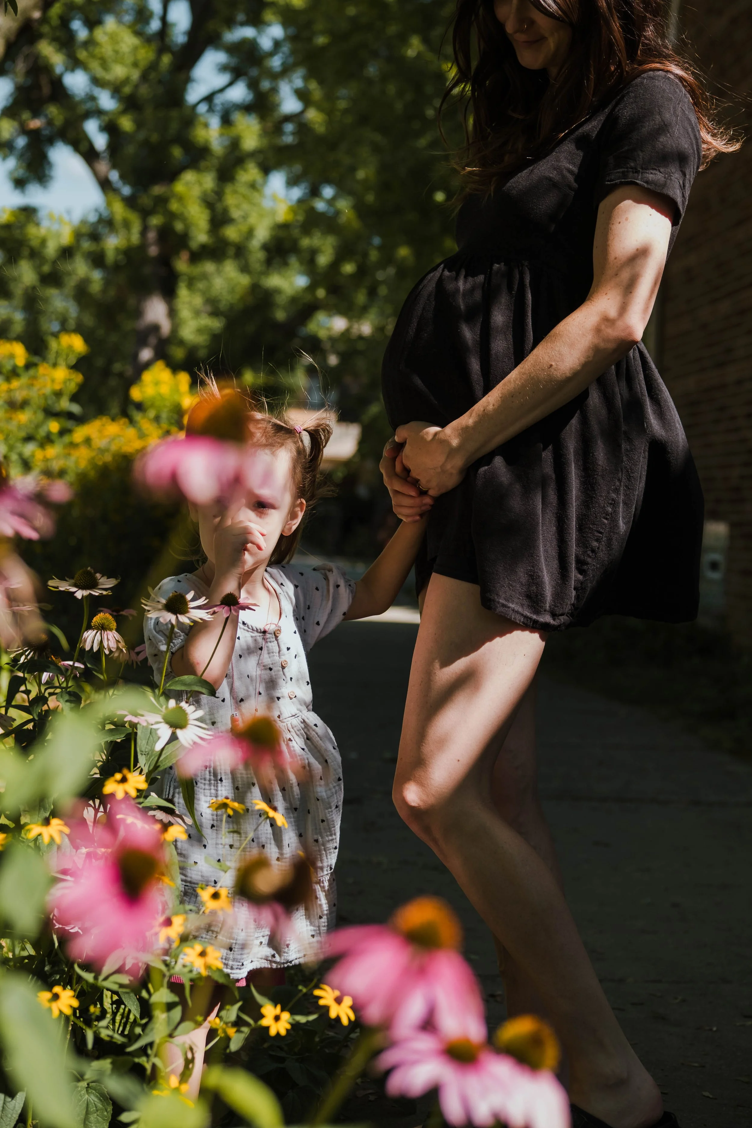 A pregnant woman wearing a black dress holding her belly while holding hands with a young girl in a garden with colorful flowers and green trees.