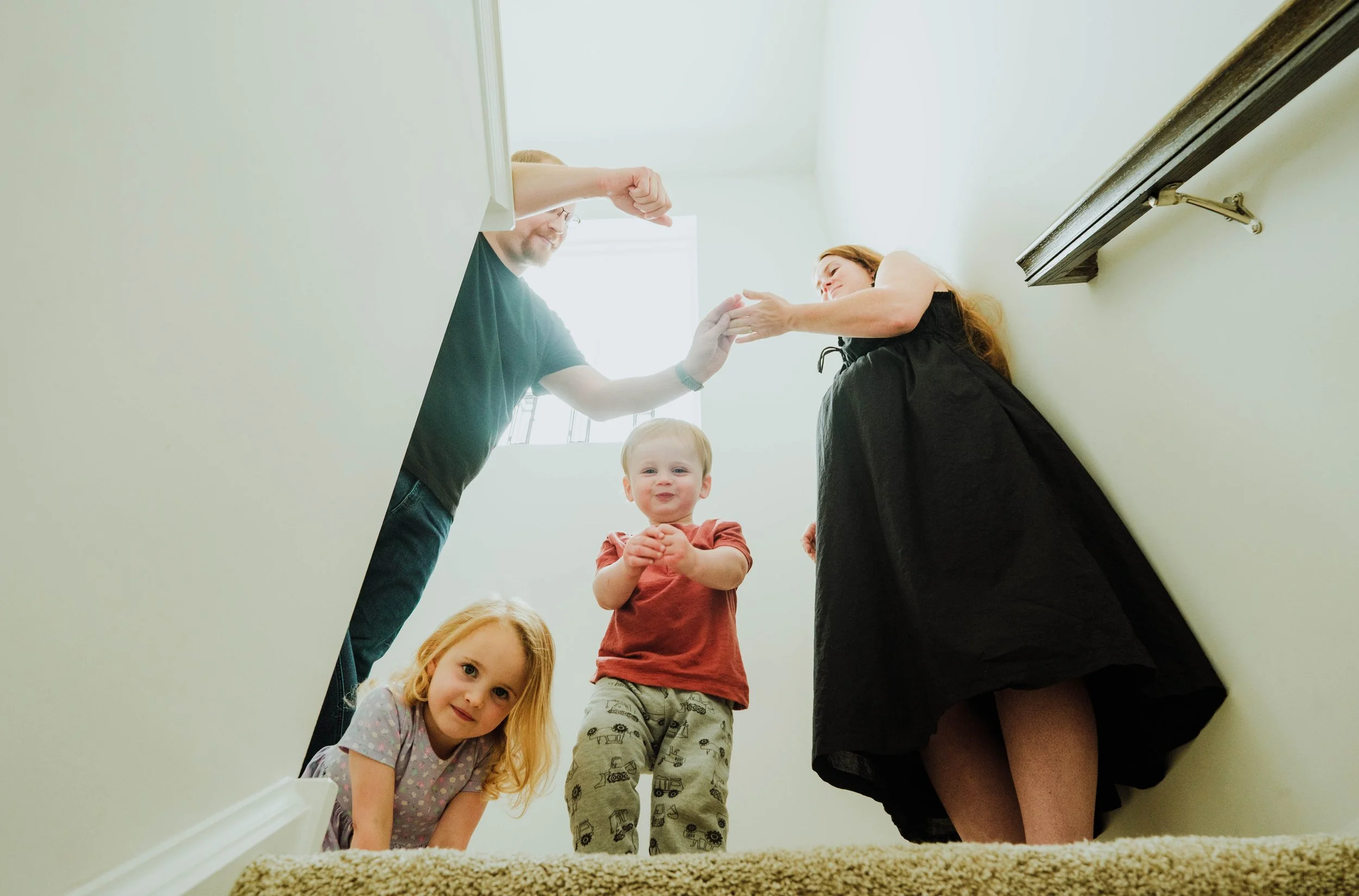 Children and adults standing on a staircase, viewed from below, engaging in play or conversation in a home.