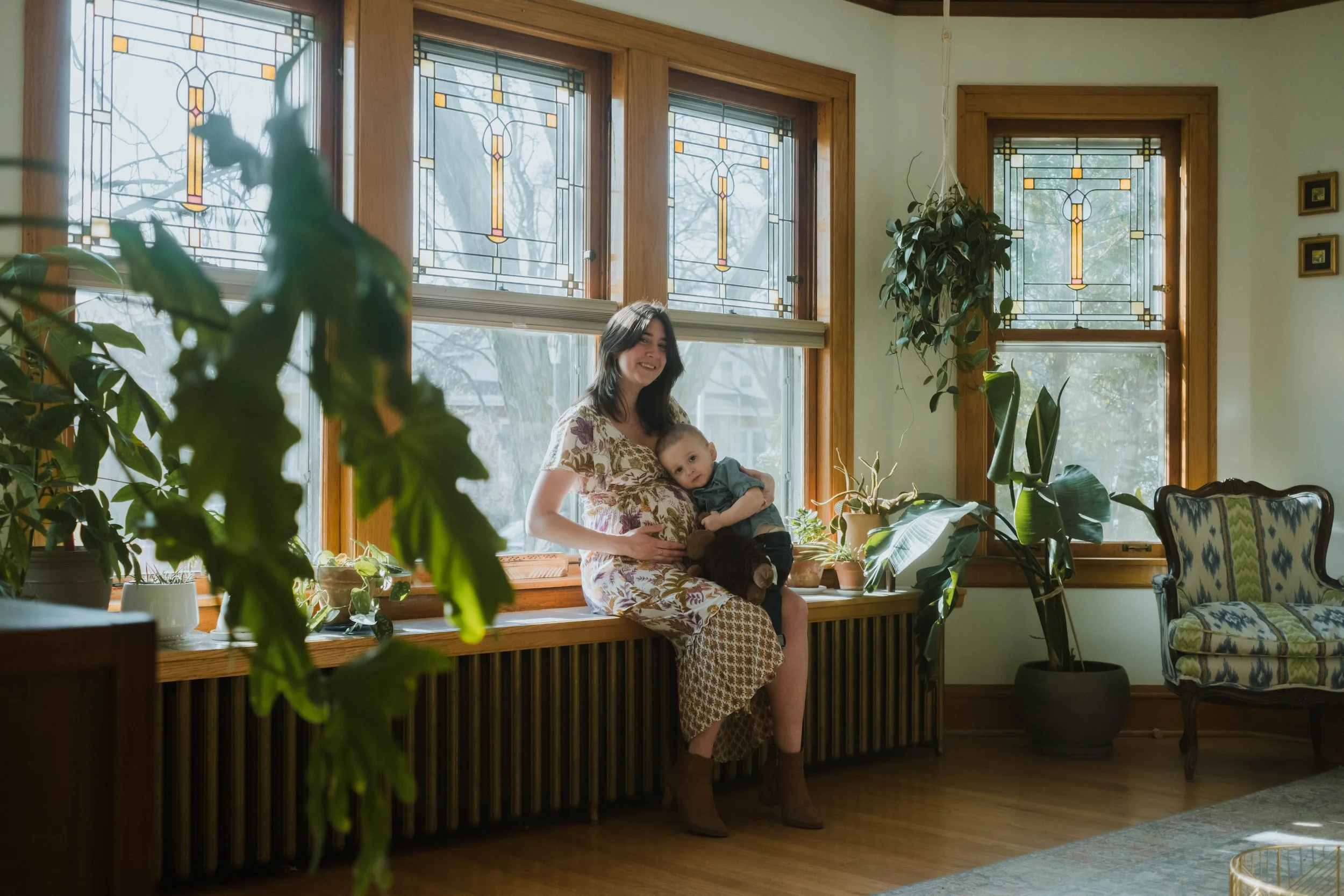A woman sitting on a wooden window seat holding a young child, inside a room with large windows featuring stained glass designs, surrounded by various green houseplants, and a patterned armchair nearby.