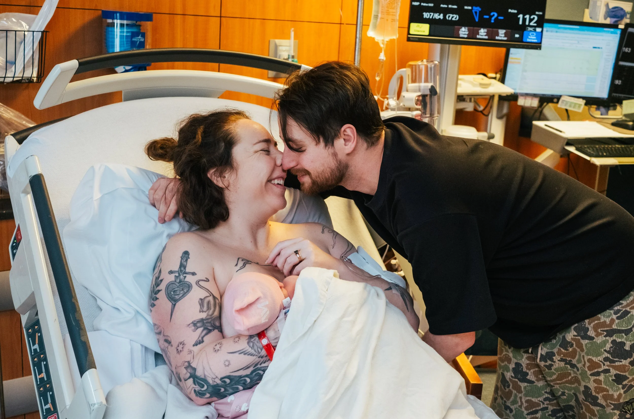 A woman in a hospital bed, smiling and touching her chest, with tattoos on her arm, and a man leaning in with his forehead touching hers, in a hospital room with medical equipment and monitors in the background.