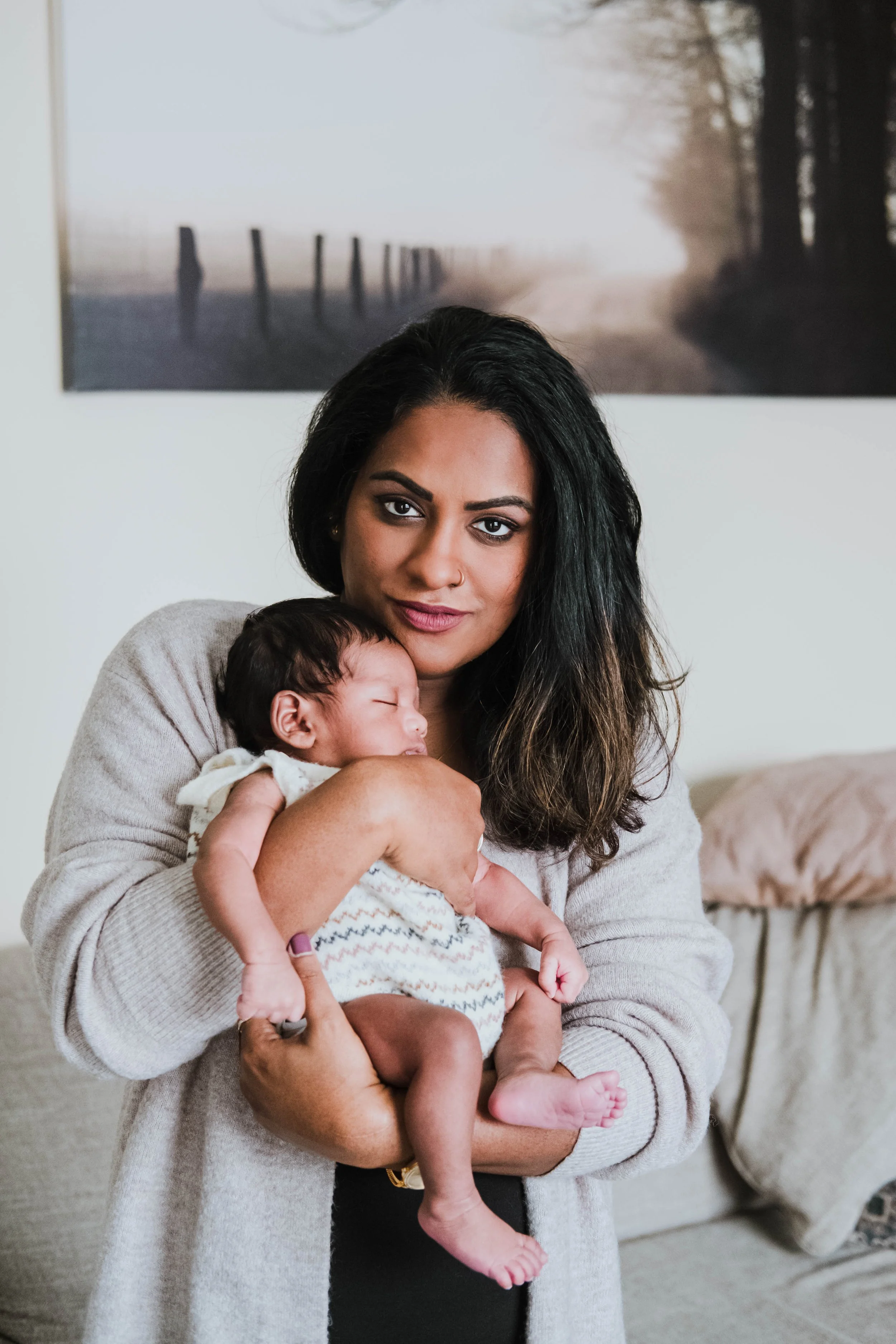 A woman with dark hair and a tan sweater holding a sleeping baby with dark hair and pajamas. They are indoors, with a photo of a foggy forest hanging on the wall behind them.